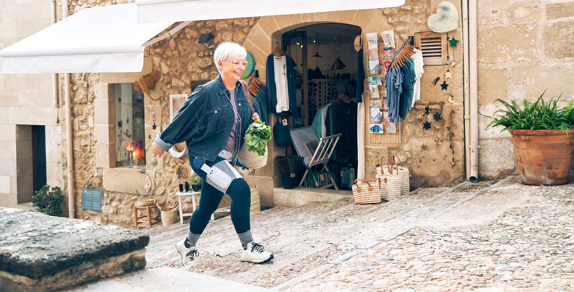 Eine ältere Frau mit kurzen weissen Haaren und Brille läuft eine gepflasterte Gasse hinauf. Sie trägt eine Jeansjacke, dunkle Leggings mit medizinischen Geräten daran und hält einen Korb mit frischem Grünzeug. Im Hintergrund befindet sich ein Geschäft mit Kleidung und Postkarten, eingerahmt von einer Steinwand mit weissen Markisen. Neben dem Eingang stehen Körbe und ein grosser Blumentopf.