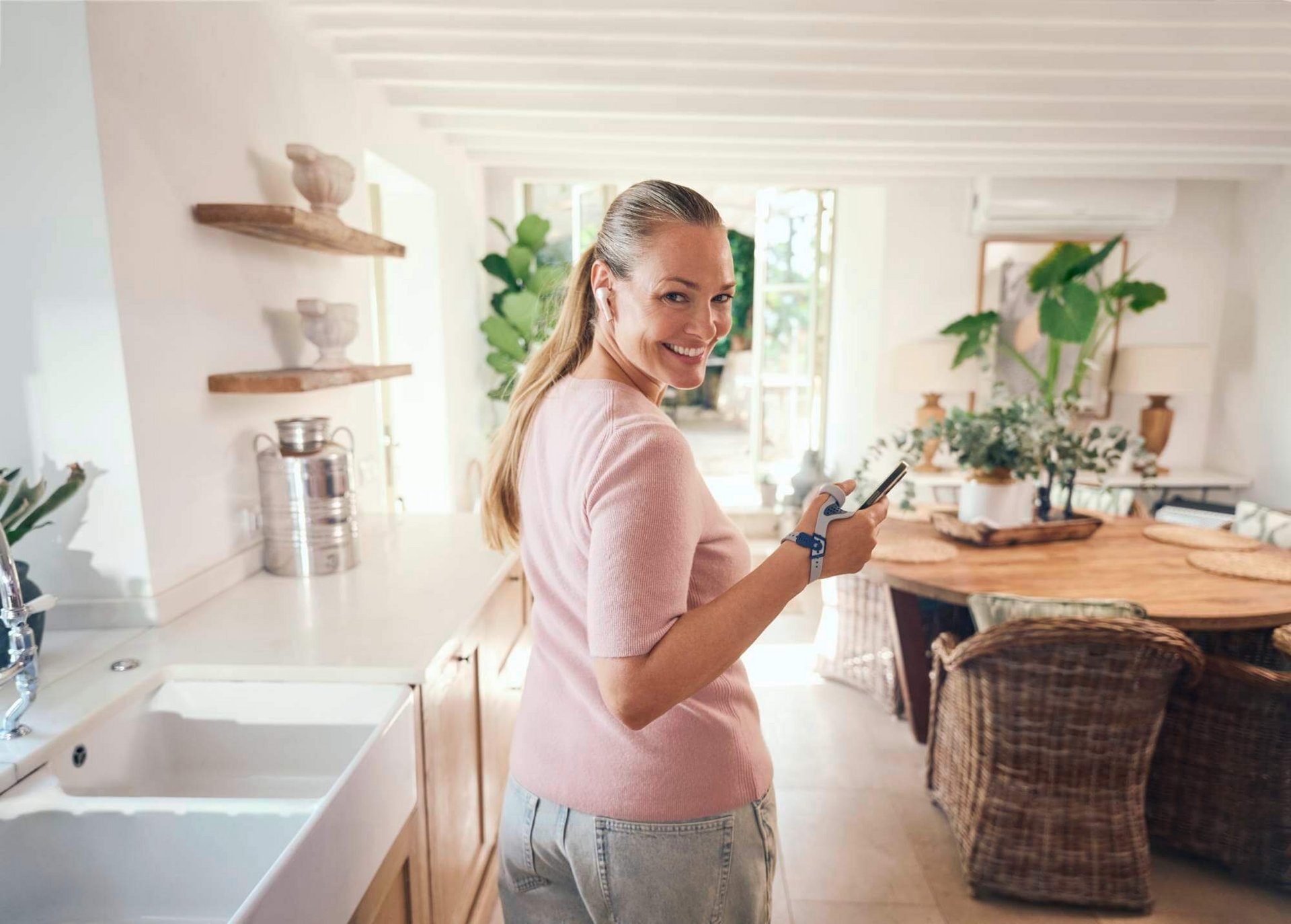 Eine lächelnde Frau mit langen blonden Haaren steht in einer hellen, modern eingerichteten Küche und hält ein Smartphone in der Hand. Sie trägt eine RhizoLoc OA Daumenorthese am rechten Handgelenk. 