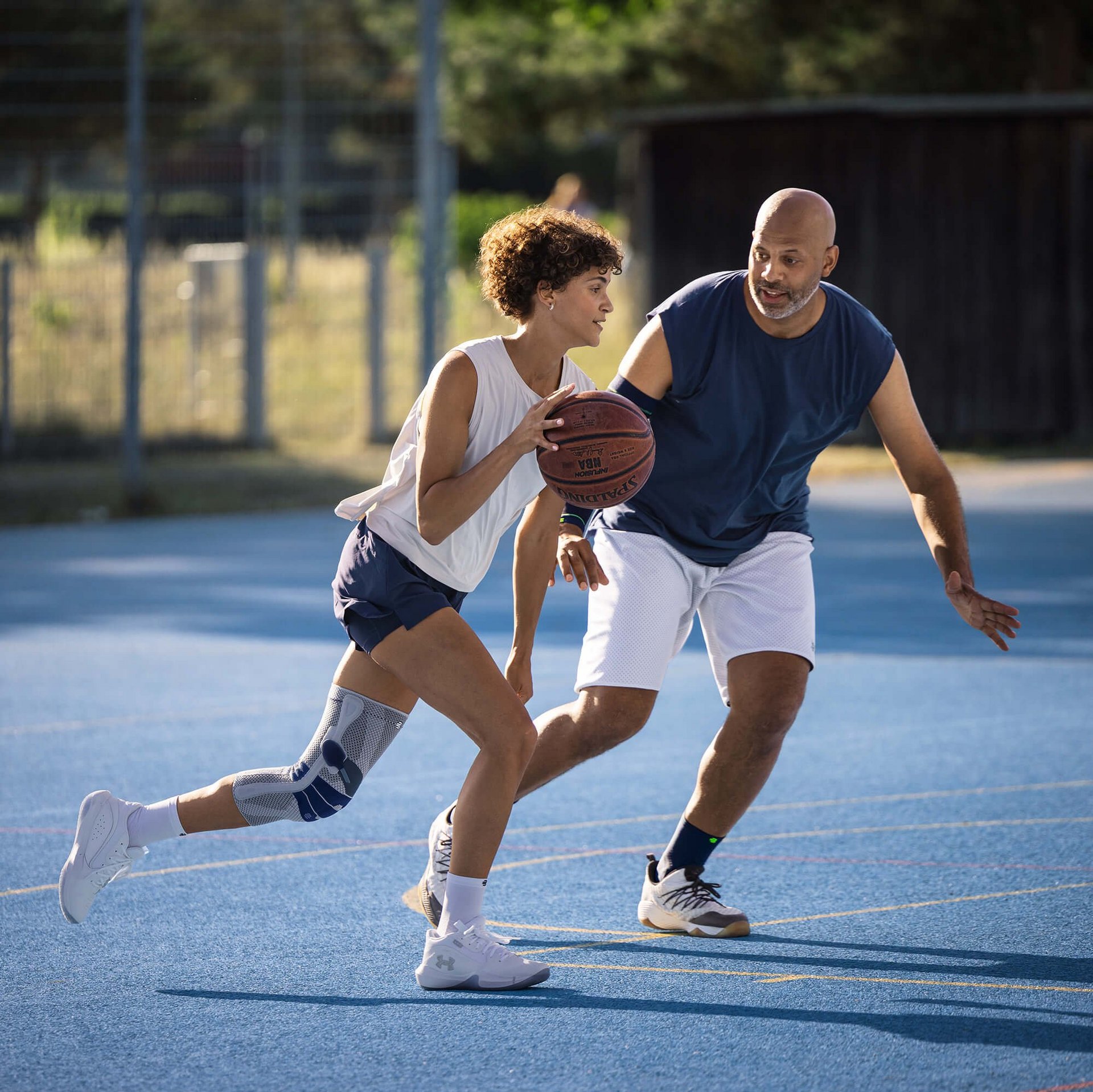 Zwei Personen spielen Basketball auf einem Aussenplatz mit blauem Bodenbelag. Die spielende Person im Vordergrund trägt eine Kniebandage am linken Bein und dribbelt den Ball, während die andere Person in Verteidigungsposition vor ihr steht. Beide sind sportlich gekleidet und konzentriert im Spielgeschehen.