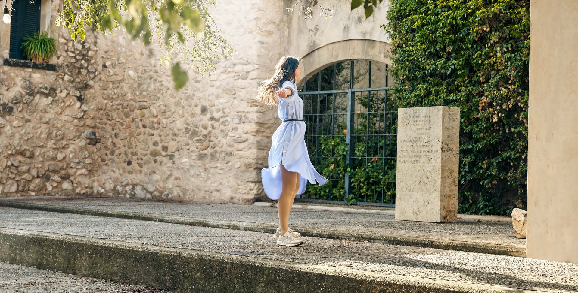 Eine junge Frau in einem fließenden hellblauen Kleid dreht sich mit ausgebreiteten Armen auf einer Steinterrasse vor einer mediterranen Steinmauer. Sie trägt dezente VenoTrain Kompressionsstrümpfe, die sich harmonisch in das Outfit einfügen.