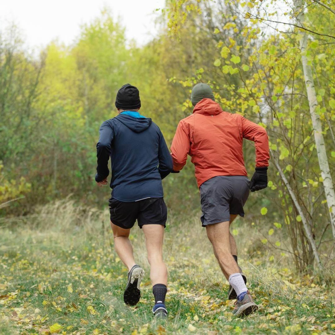 wei Männer joggen durch einen herbstlichen Wald. Beide tragen die AchilloTrain Bandage von Bauerfeind, die für Stabilität und Unterstützung sorgt.