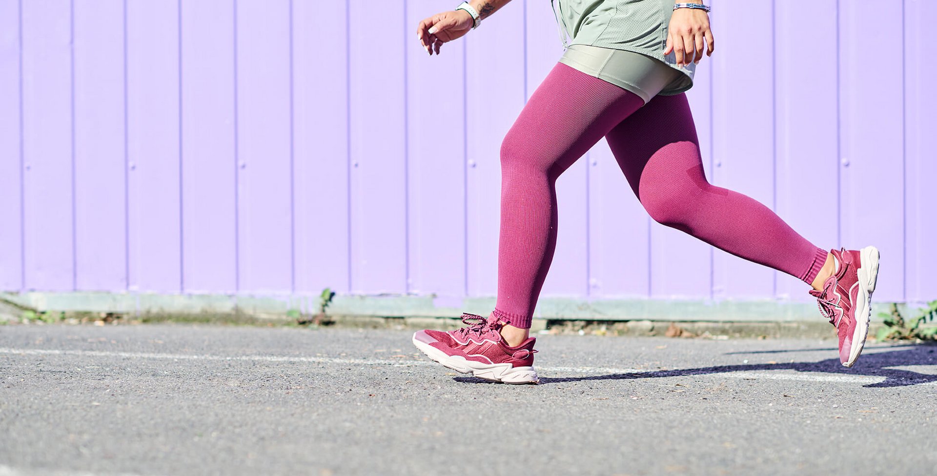 Frau in sportlicher Kleidung mit bordeauxfarbenen Strumpfhosen und passenden Turnschuhen läuft auf Asphalt vor einer lilafarbenen Holzwand. Nur der Unterkörper und ein Teil des Oberkörpers sind sichtbar.