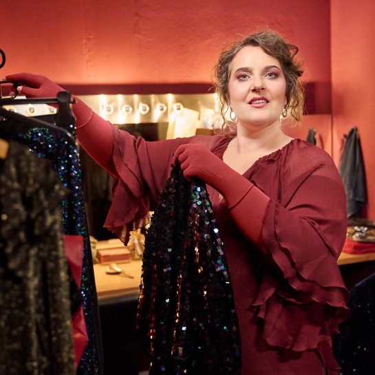 Eine Frau in einem bordeauxroten Kleid steht in einer Garderobe mit roter Wand und betrachtet ein glitzerndes, schwarzes Kleidungsstück auf einem Kleiderbügel. Im Hintergrund ist ein beleuchteter Schminktisch zu sehen.