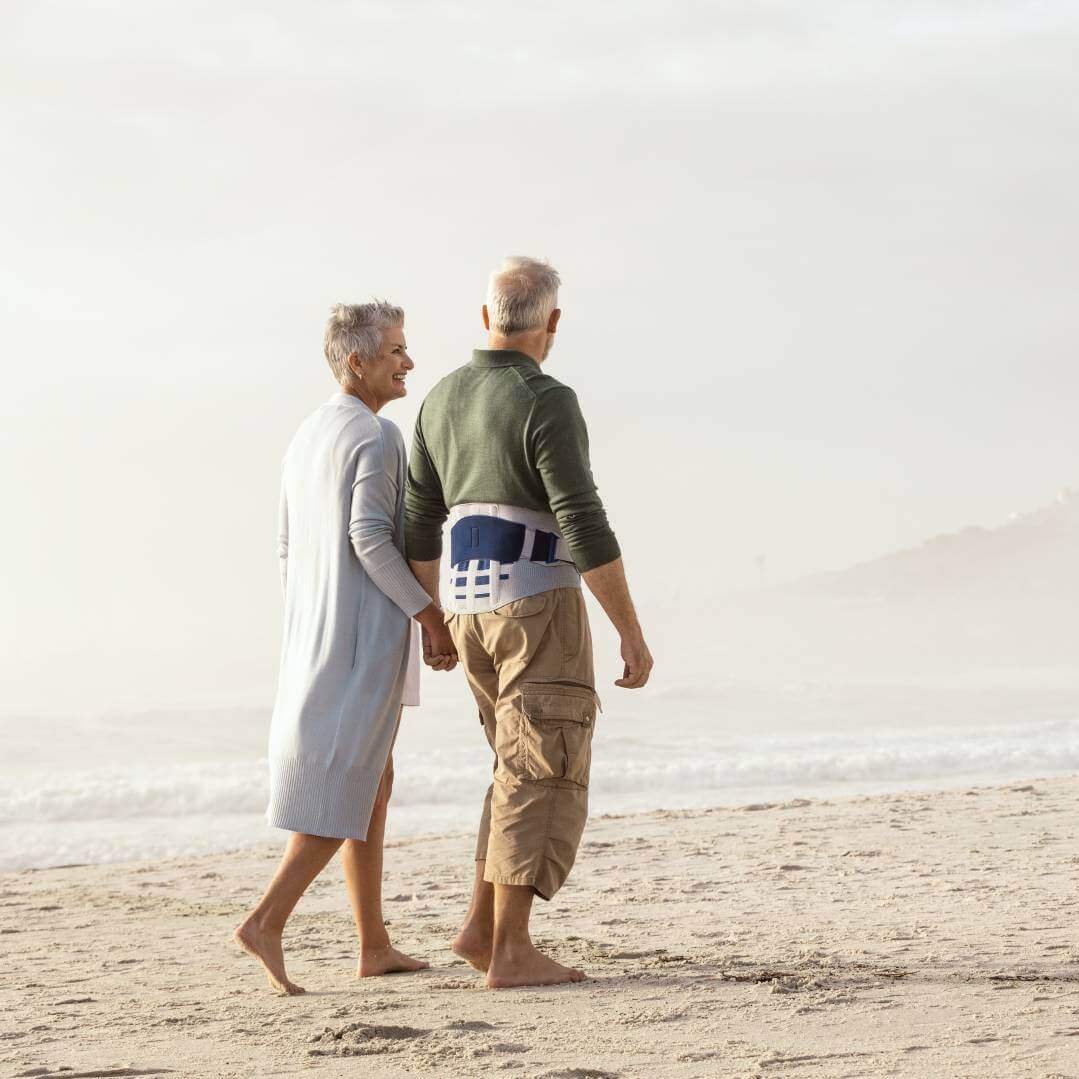 Ein älteres Paar spaziert an einem Strand entlang. Der Mann trägt dabei die LumboLoc Forte Rückenorthese von Bauerfeind.