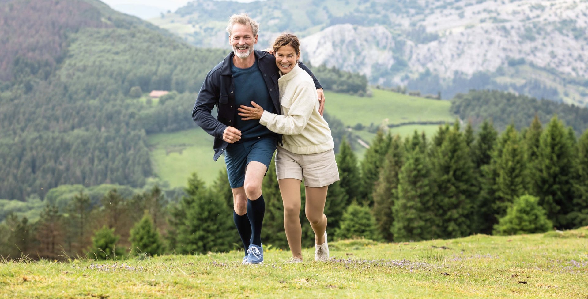 Ein Mann und eine Frau laufen Arm in Arm in einer Berglandschaft über eine Wiese. Beide tragen VenoTrain impuls Kompressionsstrümpfe von Bauerfeind. Diese helfen bei Besenreisern.