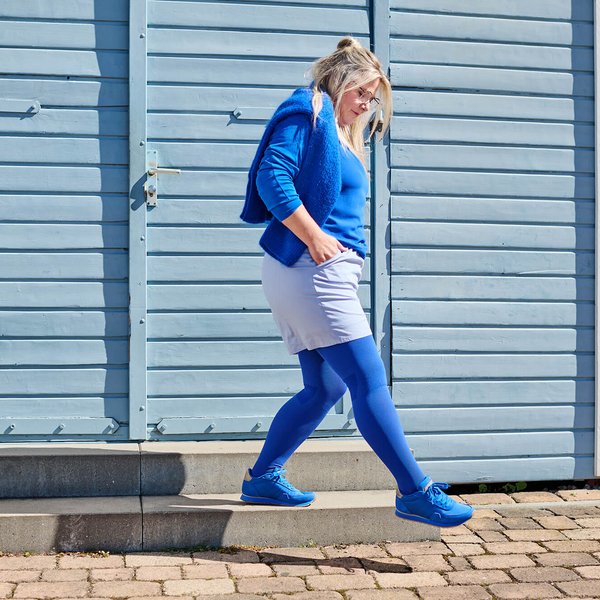 Frau mit blonden Haaren und blauer Kleidung, einschliesslich blauer Strumpfhose, Pullover und Sneakers, steigt eine kleine Betontreppe hinunter vor einer hellblauen Holzwand mit rotem Rand.