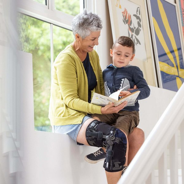 Eine ältere Frau sitzt auf einer Fensterbank und liest einem kleinen Jungen ein Buch vor. Sie trägt eine GenuTrain OA Knieorthese am rechten Bein. Im Hintergrund sind bunte gerahmte Bilder an der Wand zu sehen.