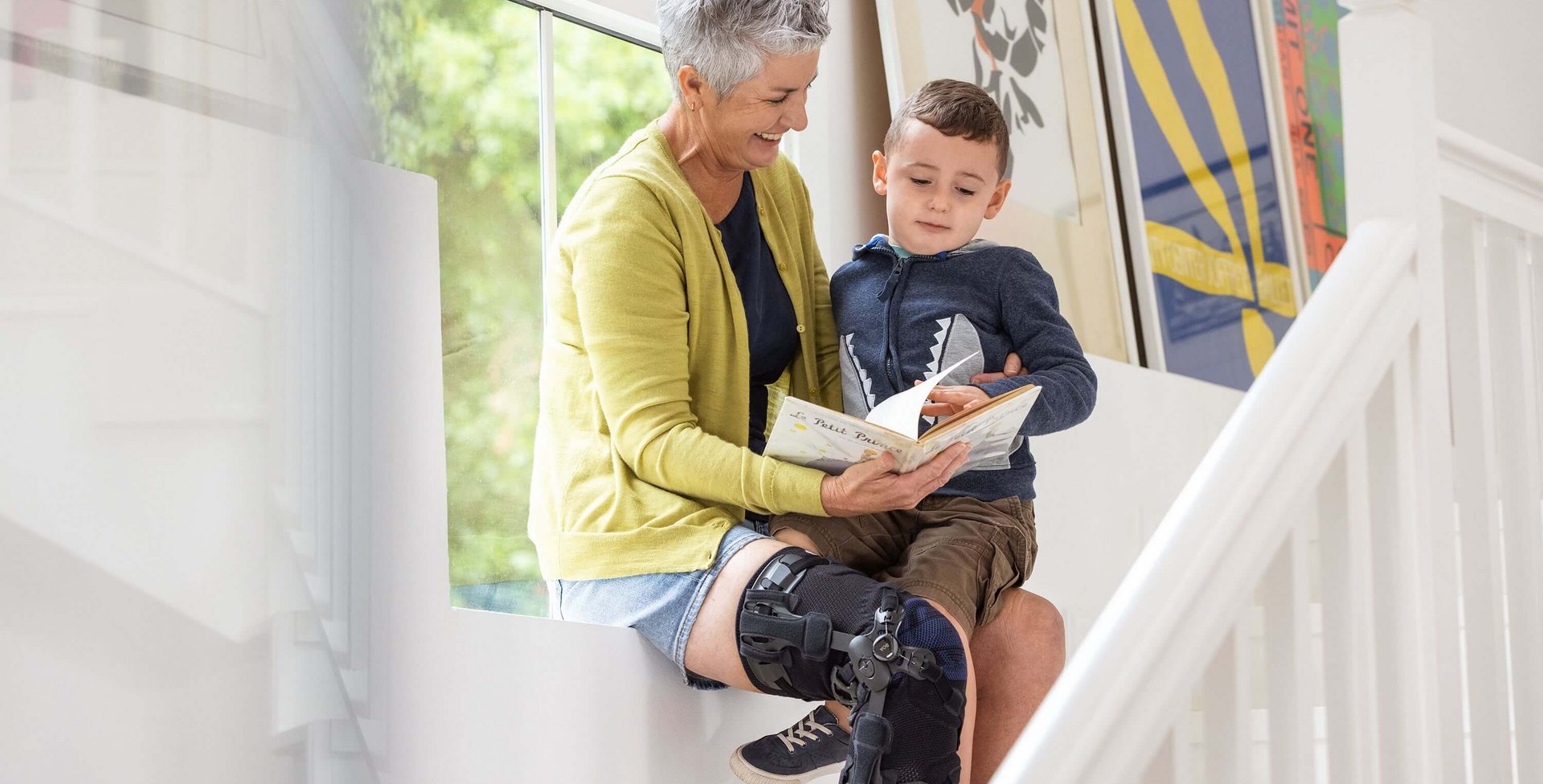 Eine ältere Frau sitzt auf einer Fensterbank und liest einem kleinen Jungen ein Buch vor. Sie trägt eine GenuTrain OA Knieorthese am rechten Bein. Im Hintergrund sind bunte gerahmte Bilder an der Wand zu sehen.