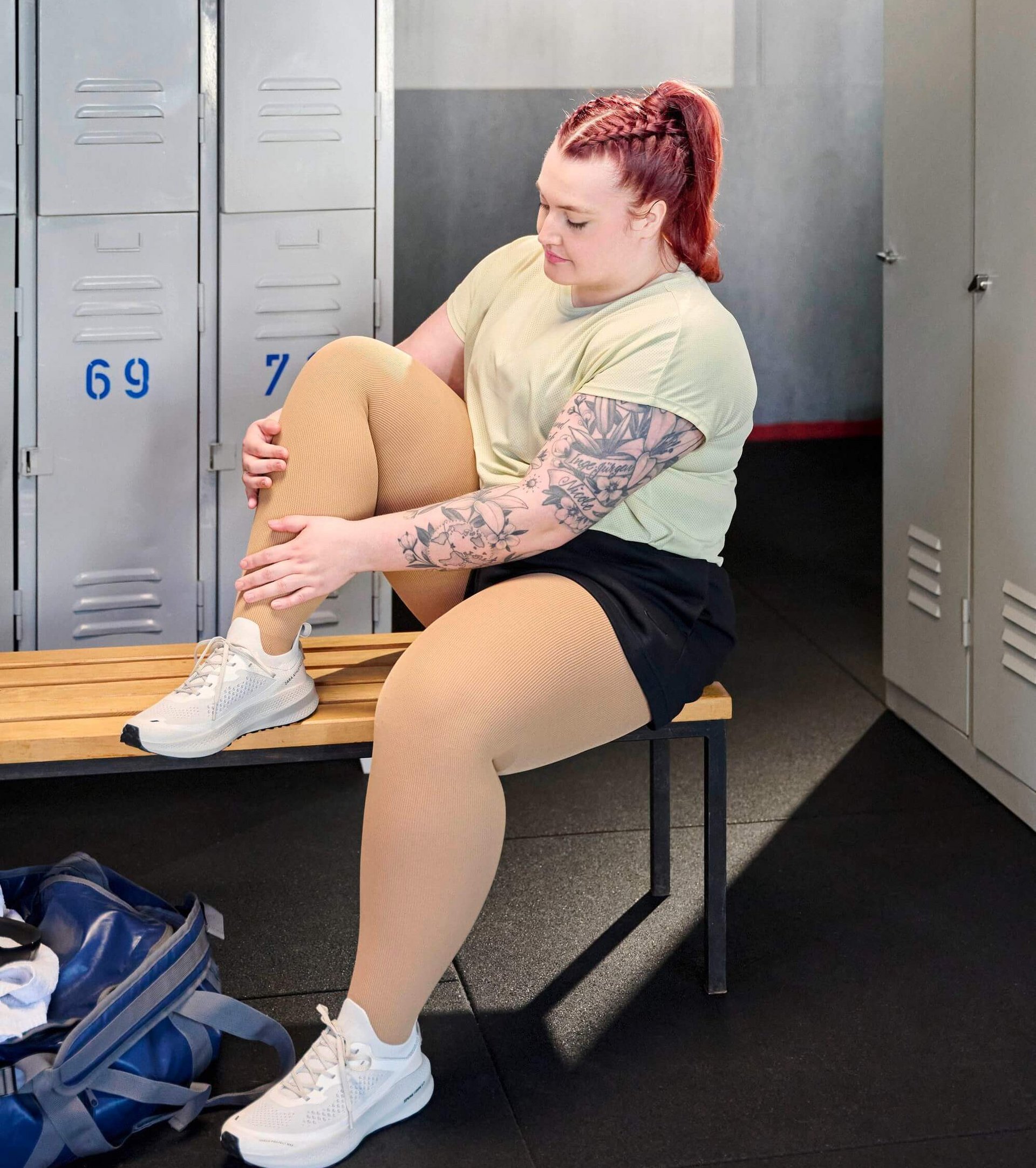 Person sitzt auf einer Holzbank in einer Umkleidekabine und hält ihr angewinkeltes Bein, das mit cremefarbenen Kompressionsstrümpfen bekleidet ist. Sie trägt ein helles Sportshirt, schwarze Shorts und weisse Sportschuhe. Im Hintergrund graue Spinde mit blauen Nummern.