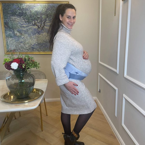 Schwangere Frau steht in einem elegant eingerichteten Raum, trägt ein helles Strickkleid, schwarze Strumpfhose, Stiefeletten und eine Schwangerschaftsorthese um den Bauch.