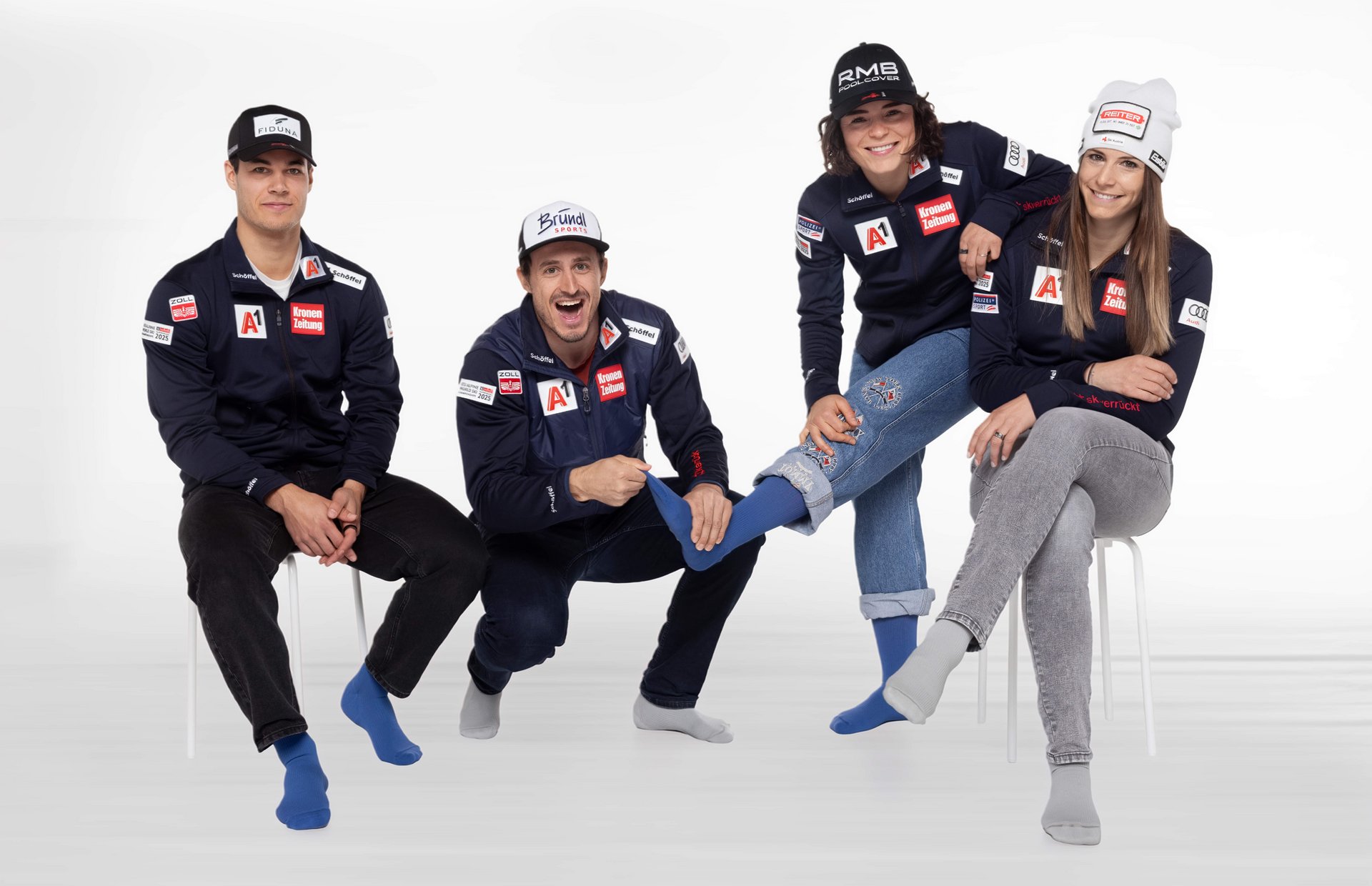 Vier Personen in sportlicher Teamkleidung mit Sponsorenlogos sitzen auf Hockern vor weissem Hintergrund und tragen bunte Socken; eine Person kniet in der Mitte und hält spielerisch den Fuss einer anderen hoch, alle wirken entspannt und gut gelaunt.