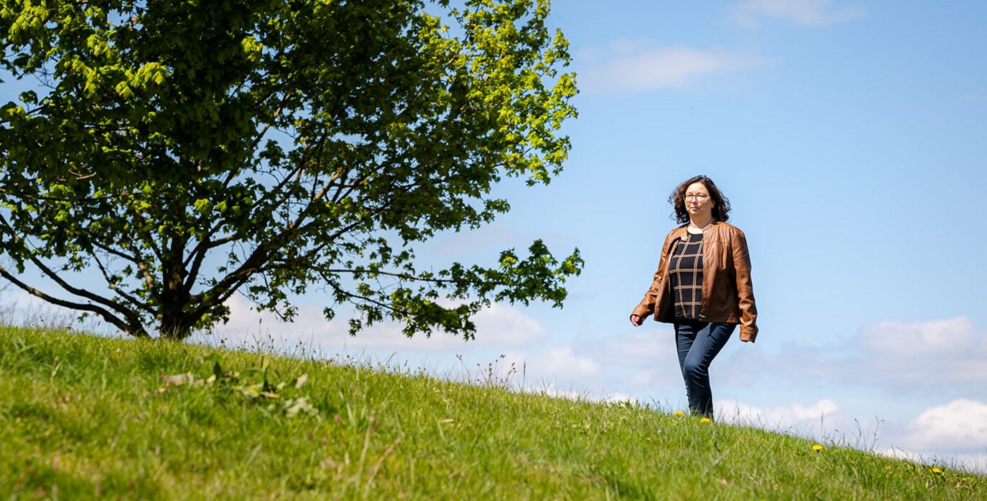Katrin, eine Lymphödempatientin, spaziert über eine grüne Wiese unter einem klaren, blauen Himmel. Im Hintergrund steht ein großer, voller Baum, der eine sommerliche und ruhige Atmosphäre vermittelt. Sie trägt eine braune Lederjacke und Jeans.
