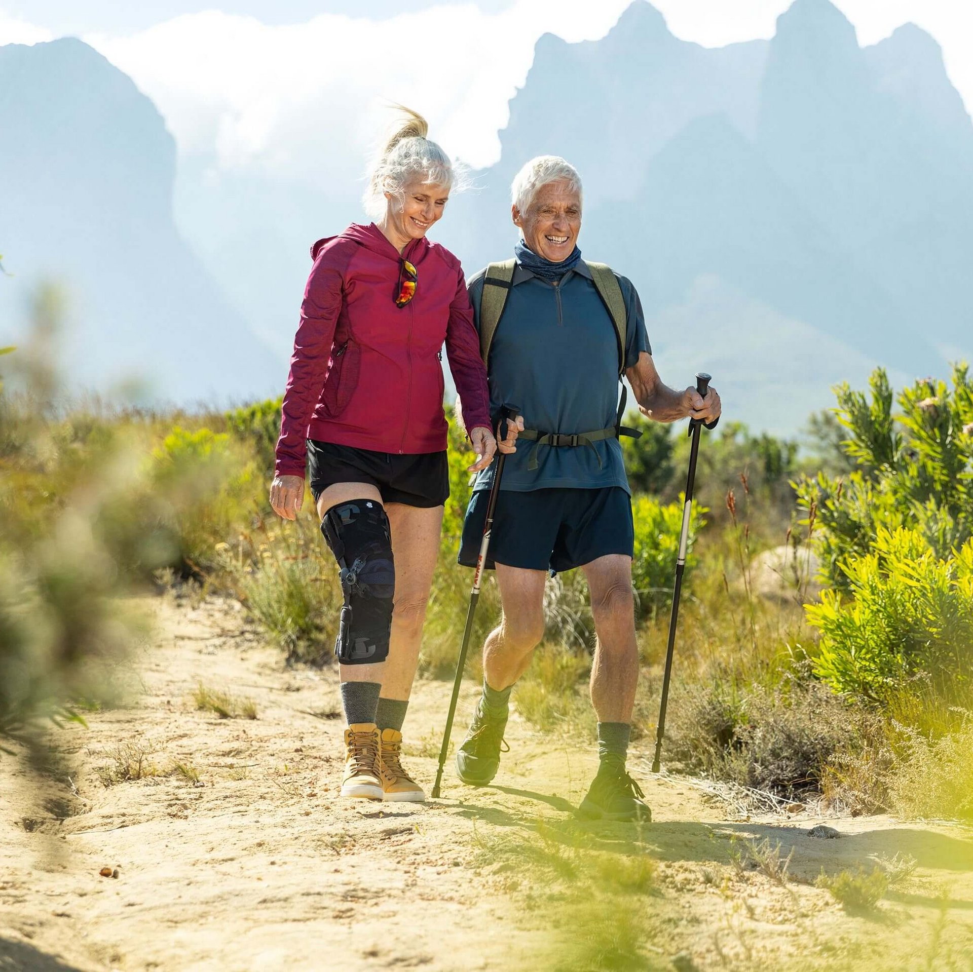 Auf dem Bild sieht man ein älteres Paar, das in einer malerischen Berglandschaft wandert. Die Frau trägt eine GenuTrain OA Orthese, die speziell für Menschen mit Arthrose entwickelt wurde, um ihr Kniegelenk zu unterstützen.