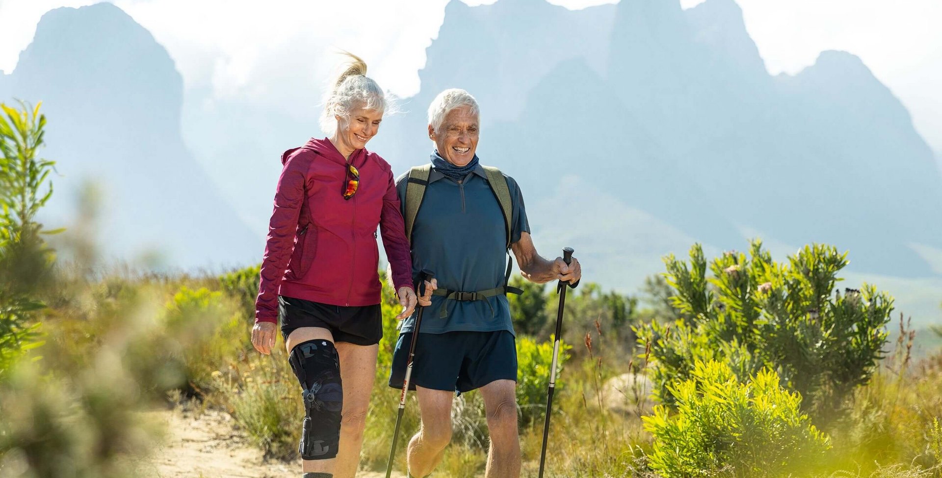  Auf dem Bild sieht man ein älteres Paar, das in einer malerischen Berglandschaft wandert. Die Frau trägt eine GenuTrain OA Orthese, die speziell für Menschen mit Arthrose entwickelt wurde, um ihr Kniegelenk zu unterstützen.