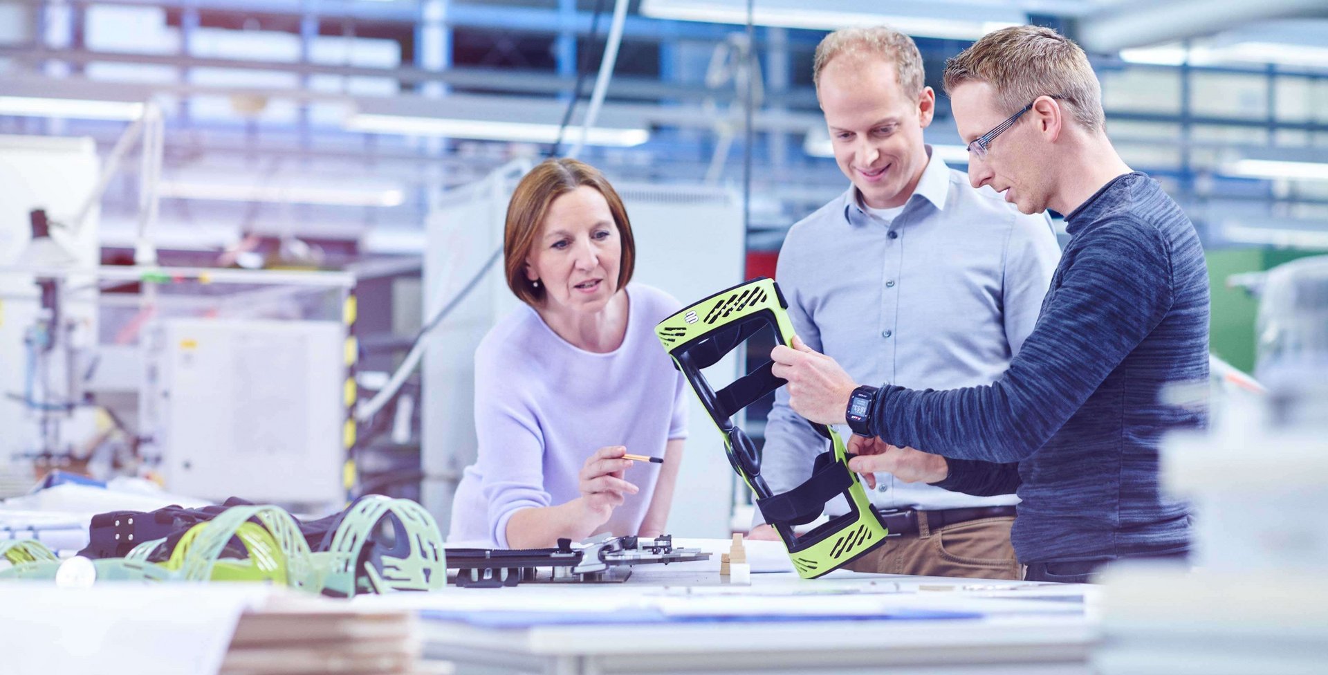 Drei Personen in Werkhalle betrachten eine grüne Rahmenorthese von Bauerfeind, eine Person hält es hoch, auf Tisch liegen technische Zeichnungen und Werkzeuge.
