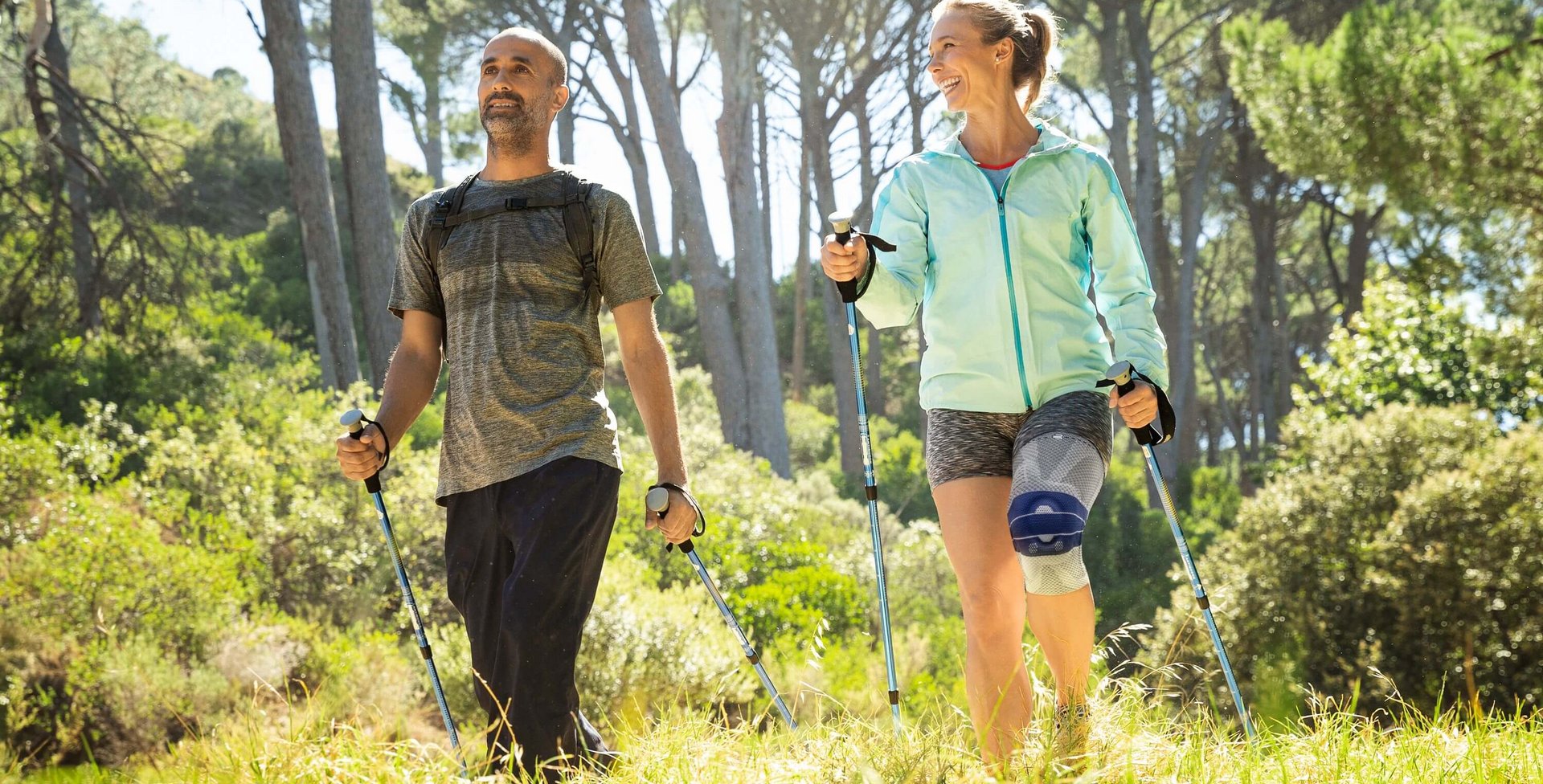 Ein Mann und eine Frau wandern mit Nordic-Walking-Stöcken durch einen sonnigen, grünen Wald. Beide tragen sportliche Kleidung, und die Frau trägt eine Kniebandage. Sie lächeln sich an und wirken entspannt.