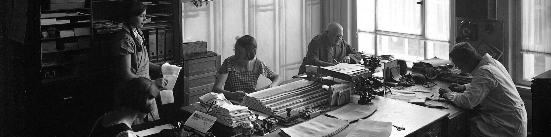Schwarz-weiss-Foto eines alten Büros mit fünf Personen, die an einem grossen Tisch arbeiten. Es herrscht eine geschäftige Atmosphäre mit Schreibmaschinen, Papierstapeln und Hängelampen. Im Hintergrund sind grosse Fenster und ein Aktenschrank mit zahlreichen Schachteln zu sehen.