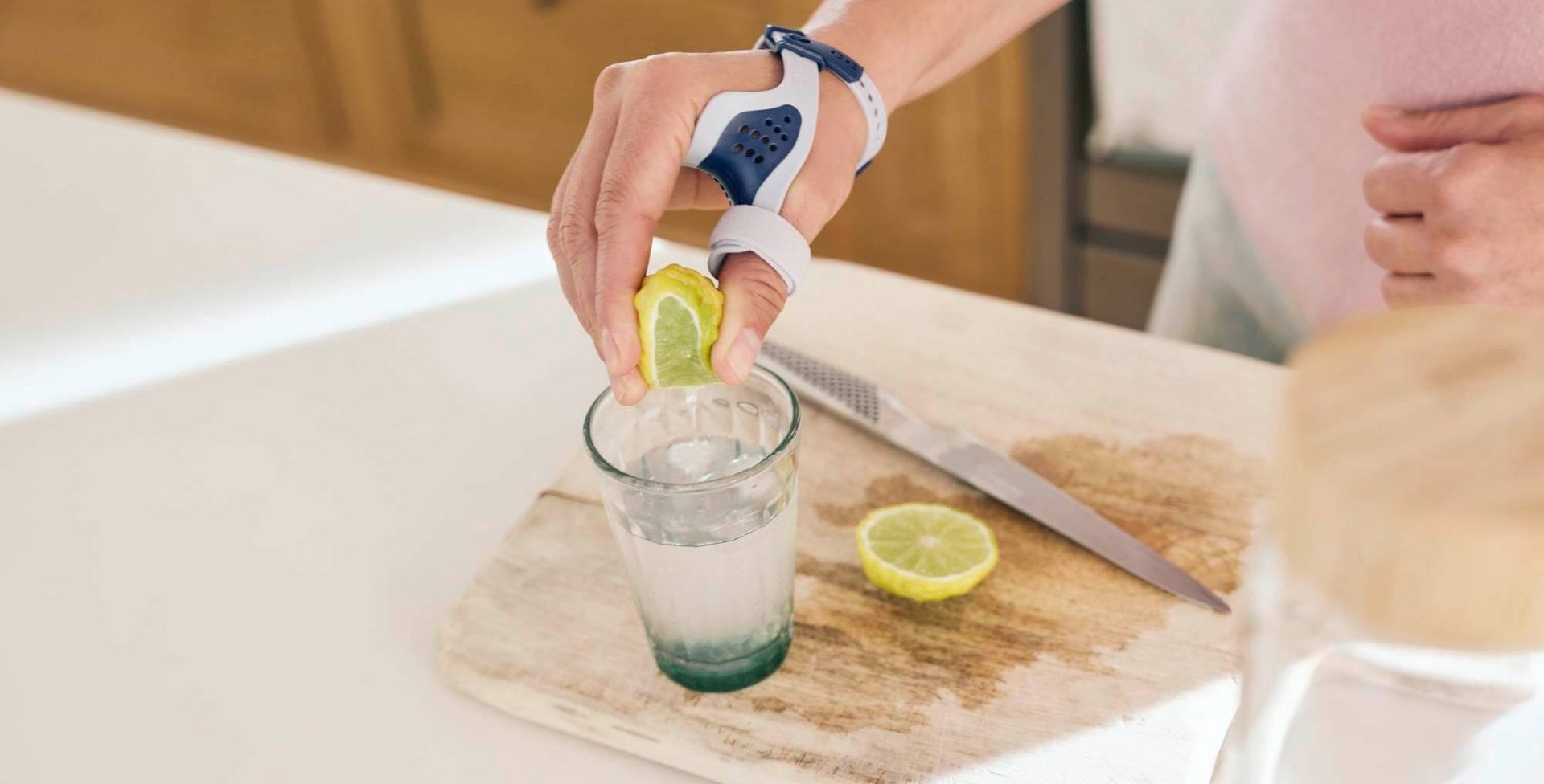 Nahaufnahme einer Hand, die eine Zitronenhälfte über einem Glas Wasser auspresst. Die Person trägt eine RhizoLoc OA Daumenorthese am rechten Daumen. Auf dem hölzernen Schneidebrett liegen ein Messer und eine weitere Zitronenscheibe. 