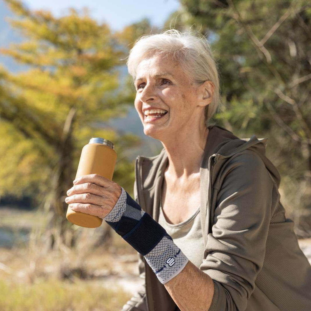 Auf dem Bild ist eine ältere Frau mit Getränk an einem See zu sehen, die beim Wandern eine ManuTrain Handgelenkbandage trägt, um Schmerzen im Handgelenk zu lindern.