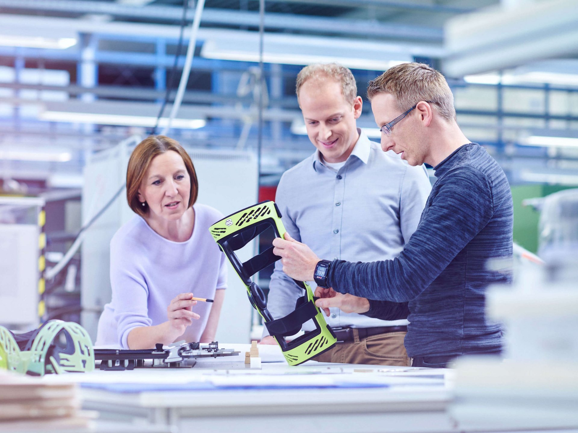 Drei Personen stehen an einem großen Tisch in der Entwicklungsabteilung und schauen auf die Rahmenorthese SecuTec Genu. Sie betrachten und besprechen ein hellgrünes technisches Bauteil, vermutlich ein Gehäuse oder eine Halterung, das der Mann rechts in den Händen hält. Links auf dem Tisch liegen weitere Bauteile und Pläne.
