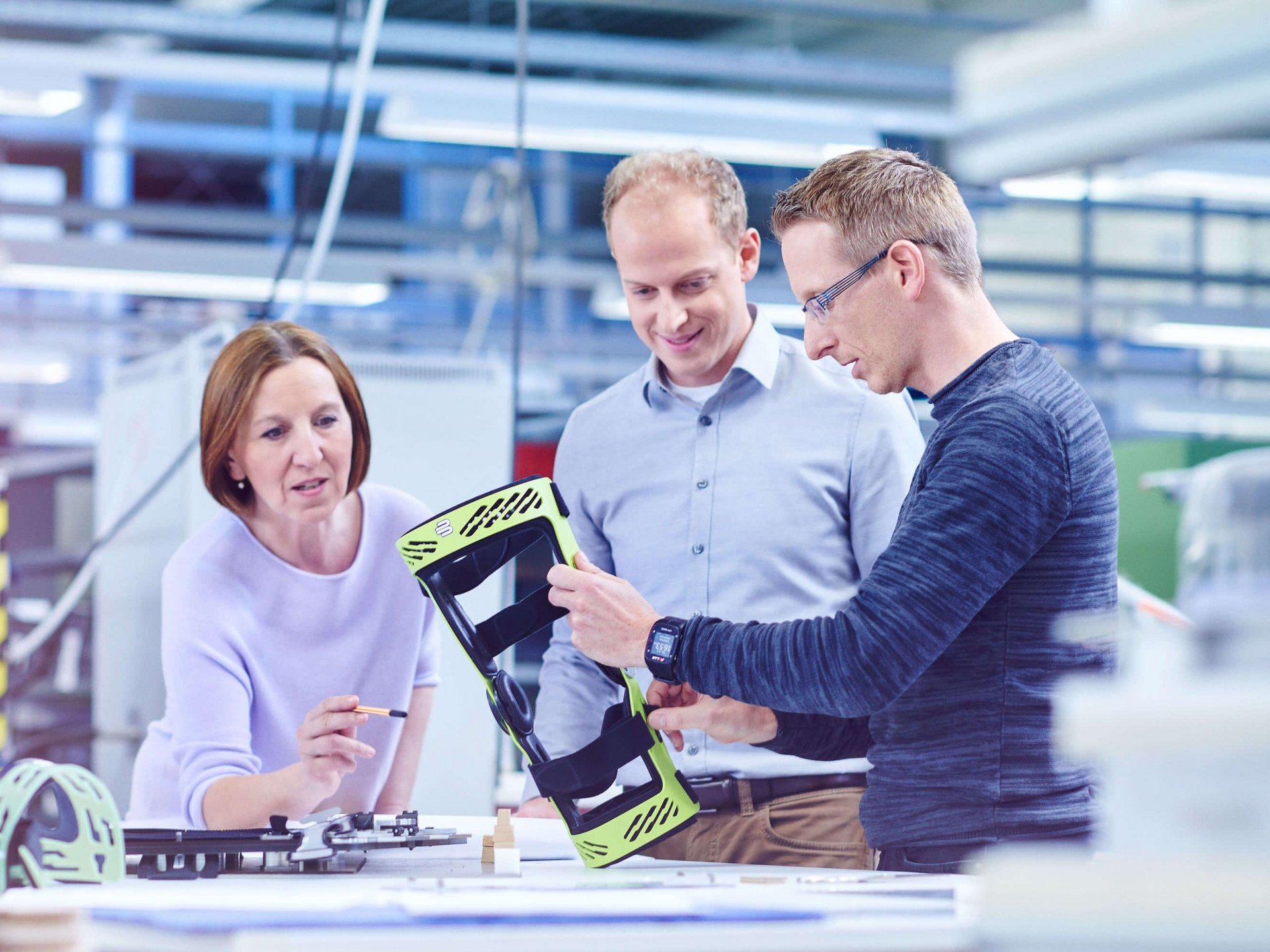 Drei Personen stehen an einem großen Tisch in der Entwicklungsabteilung und schauen auf die Rahmenorthese SecuTec Genu. Sie betrachten und besprechen ein hellgrünes technisches Bauteil, vermutlich ein Gehäuse oder eine Halterung, das der Mann rechts in den Händen hält. Links auf dem Tisch liegen weitere Bauteile und Pläne.