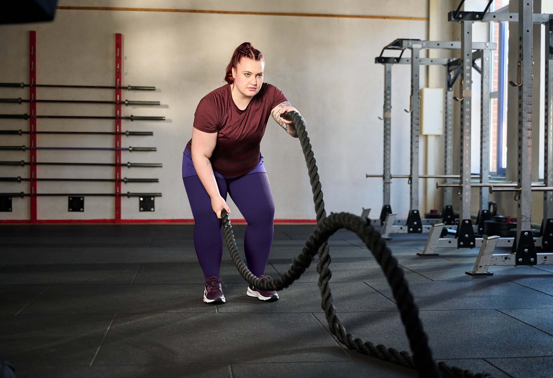 Eine Frau trainiert in einem Fitnessstudio mit Battle Ropes. Sie trägt ein bordeauxfarbenes T-Shirt, lila Leggings und Sportschuhe. Im Hintergrund sind Hanteln an der Wand sowie Krafttrainingsgeräte zu sehen.
