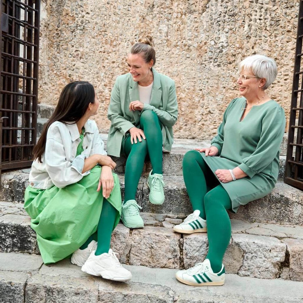 Drei Frauen sitzen auf historischen Steinstufen vor einer rustikalen Steinmauer und unterhalten sich lachend. Sie tragen Outfits in verschiedenen Grüntönen, ergänzt durch VenoTrain Kompressionsstrümpfe in der Modefarbe 'Irish Green'.