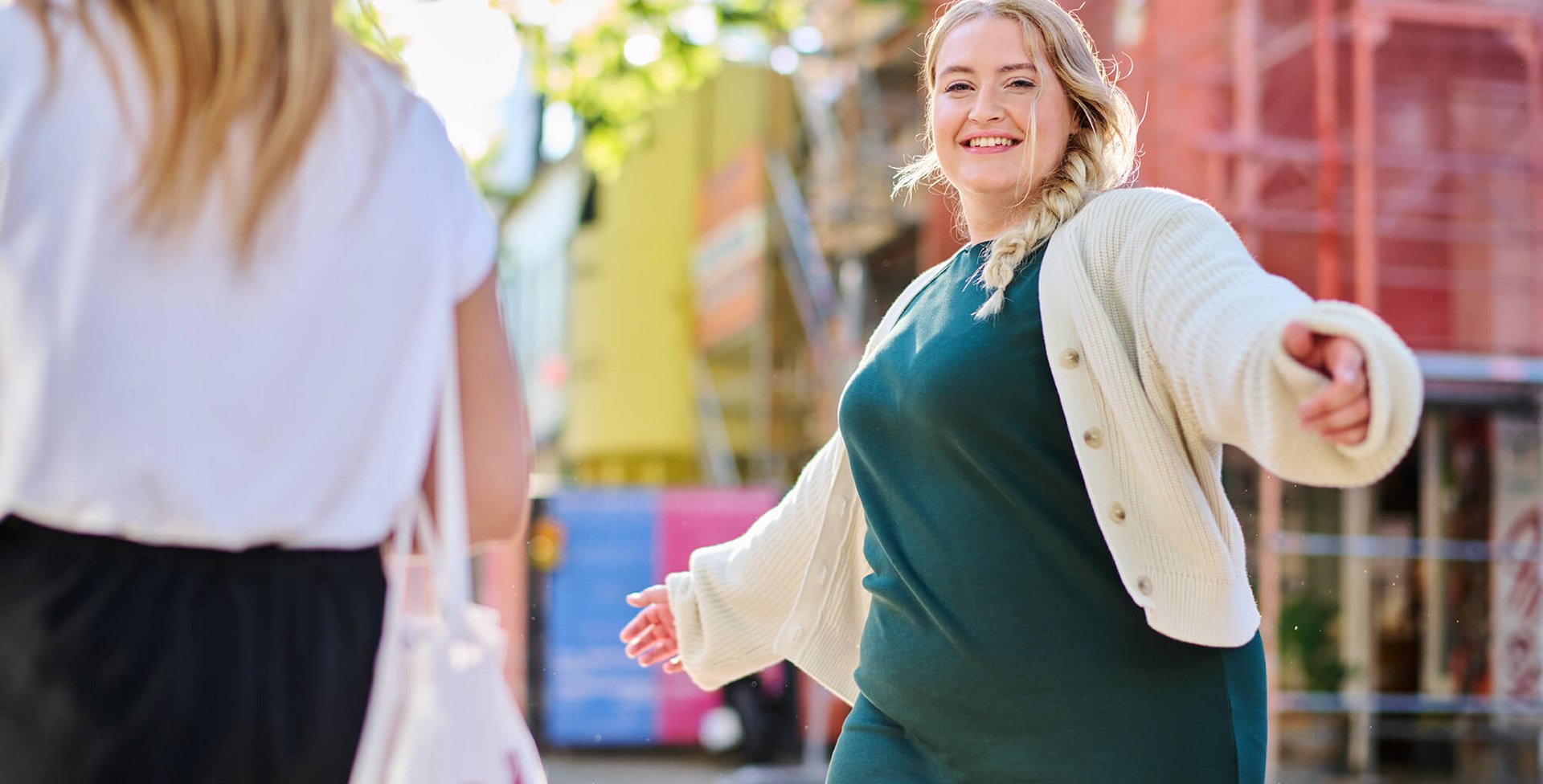 Zwei Frauen begegnen sich auf einer sonnigen Strasse in einer Stadt. Eine Frau mit blondem Zopf, grünem Kleid und weisser Strickjacke breitet die Arme freudig aus und lächelt der anderen Frau entgegen. Im Hintergrund sind bunte Baugerüste und Gebäude zu sehen.