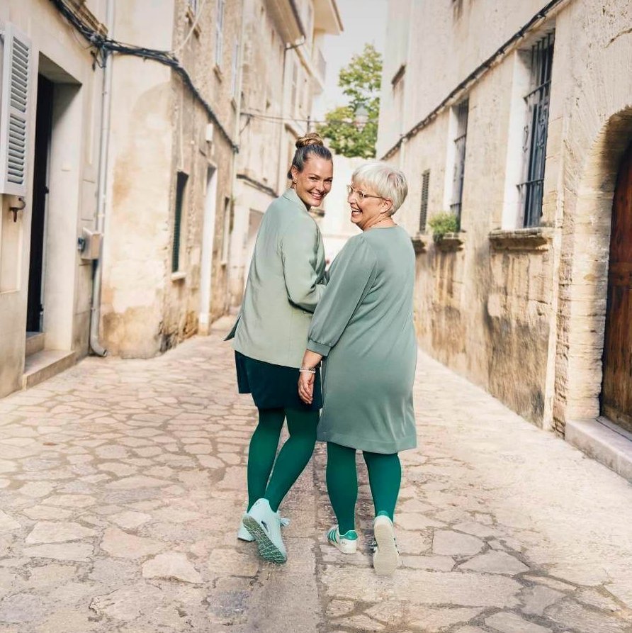 Zwei Frauen, eine jüngere und eine ältere, spazieren lachend Hand in Hand durch eine charmante, enge Altstadtgasse. Beide tragen farblich abgestimmte Outfits in Grün mit passender Kompression und Sneakern.