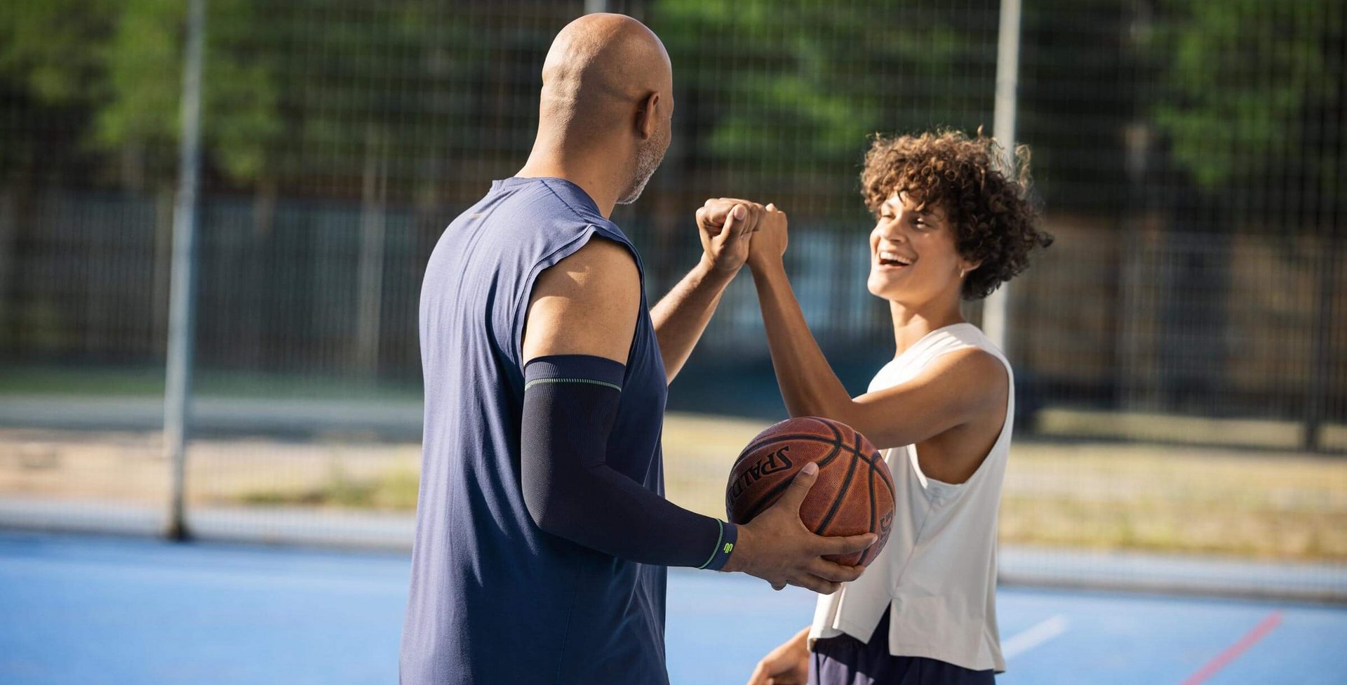 Zwei Basketballspieler auf einem blauen Aussenplatz geben sich lächelnd einen Fauststoss. Die Person links, grösser und mit Glatze, hält einen Basketball und trägt ein ärmelloses Trikot. Die Person rechts hat lockige Haare, trägt ein weisses Sporttop und wirkt fröhlich. Im Hintergrund ist ein hoher Zaun mit Bäumen sichtbar.