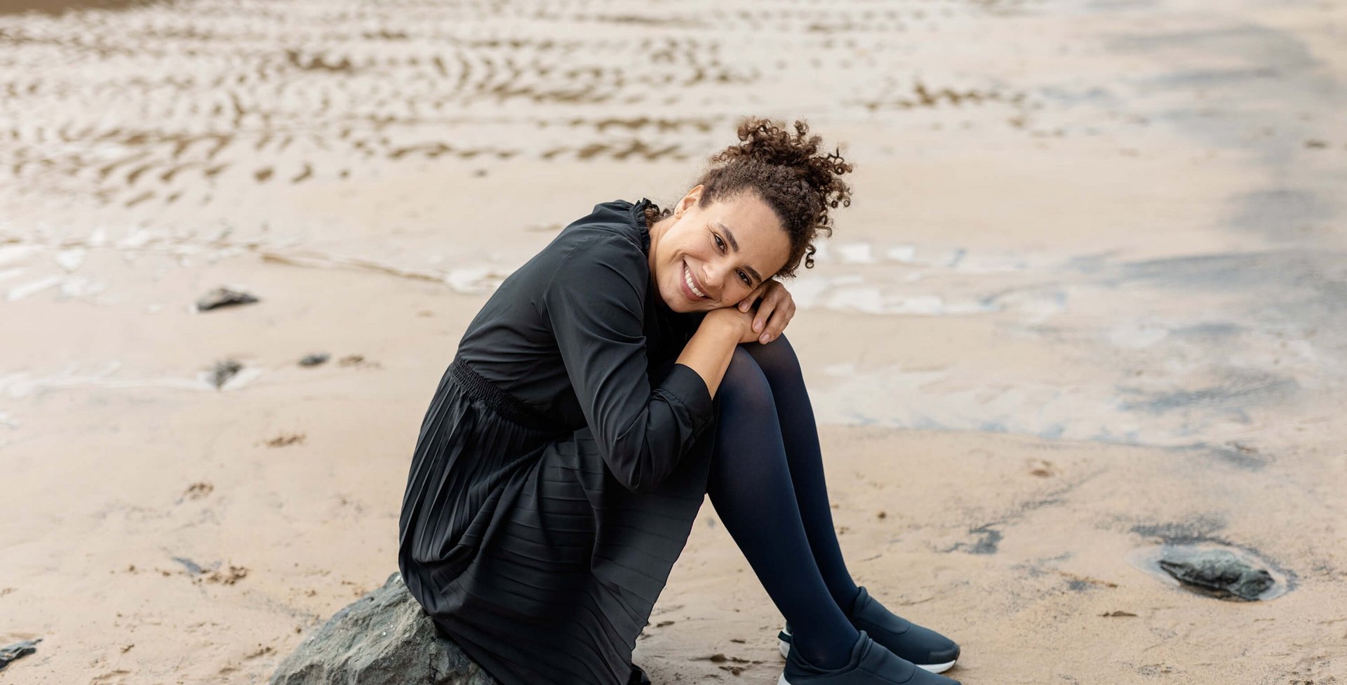 Das Bild zeigt eine Frau die auf einem kleinen Felsen am Strand sitzt. Sie trägt VenoTrain soft Kompressionsstrümpfe von Bauerfeind.