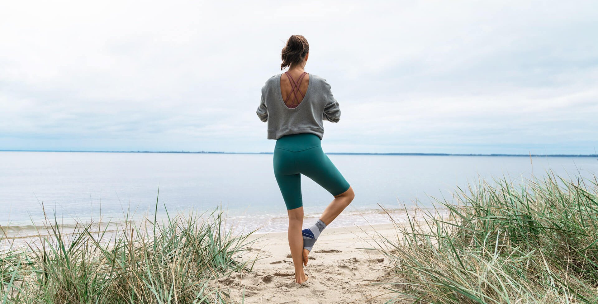 Das Bild zeigt eine Frau, die am Strand eine Yoga-Pose einnimmt. Sie trägt die MalleoTrain Sprunggelenkbandage von Bauerfeind, um ihr Sprunggelenk zu stabilisieren. Diese kommt u.a. bei einer Sprunggelenksdistorsion zum Einsatz.