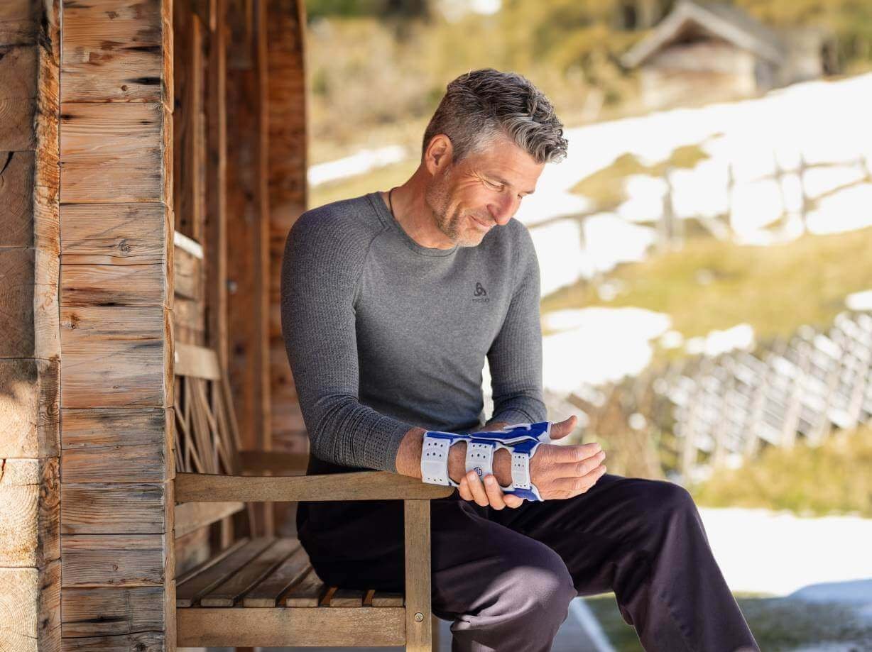 Auf dem Bild ist ein Mann zu sehen, der auf einer Holzbank vor einer Hütte sitzt. Er fasst sich an die rechte Hand an der er eine ManuLoc Rhizo von Bauerfeind trägt.