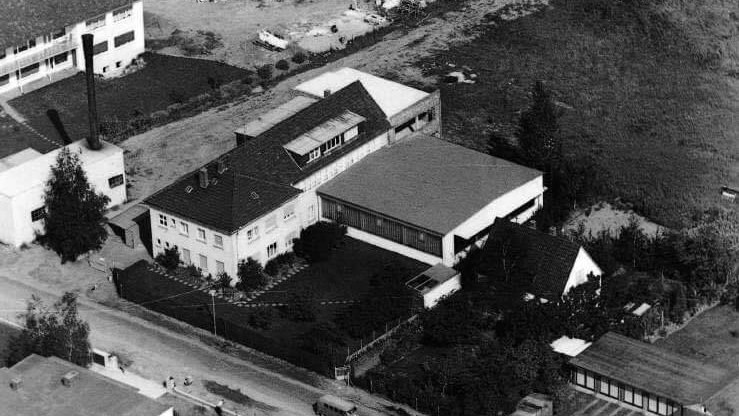 Schwarz-weiss Luftaufnahme eines Industriegebiets mit mehreren Gebäuden, darunter ein grösseres Wohnhaus mit angrenzender Halle und umliegenden Werkstätten. Im Vordergrund verläuft eine Strasse. Im Hintergrund sind unbebaute Flächen und Baumgruppen zu sehen.