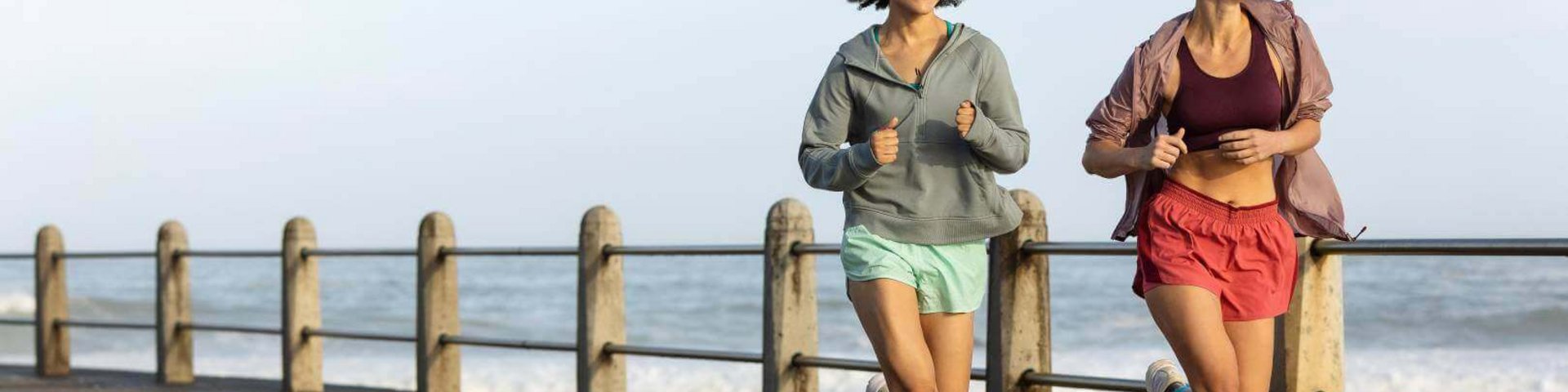 Zwei Frauen joggen lachend auf einer Promenade am Meer entlang. Die linke Frau trägt eine graue Trainingsjacke, hellgrüne Shorts und dunkelrote Kompressionssocken.