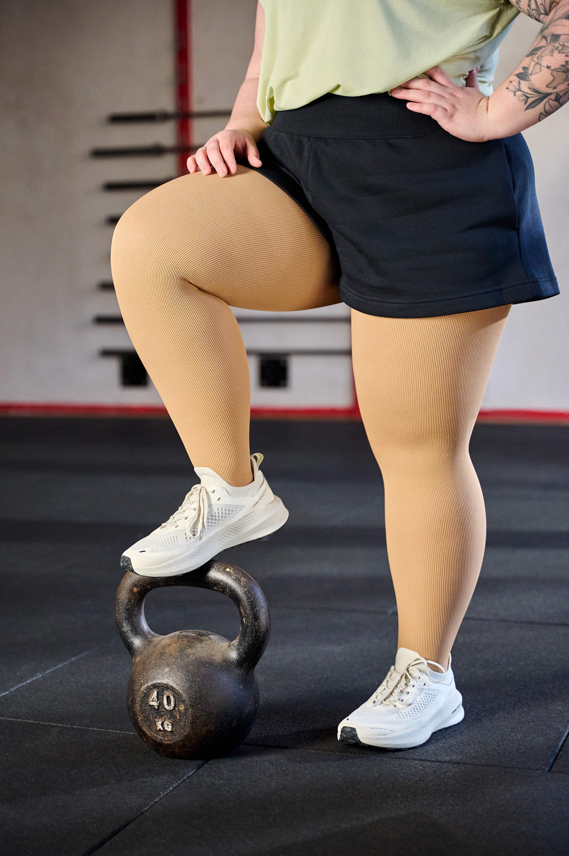 Nahaufnahme der Beine einer Person in cremefarbenen Kompressionsstrümpfen und weissen Sportschuhen. Die Person trägt eine schwarze Shorts und hat ein Bein auf eine 40-kg-Kettlebell in einem Fitnessstudio gestellt.