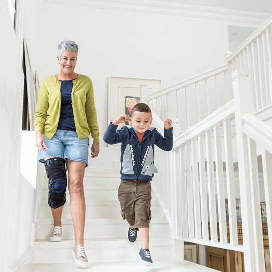 Das Bild zeigt eine Dame mit ihrem Enkel beim Herunterlaufen einer Treppe. Zur Unterstützung ihres Knies trägt die Frau eine Gonarthrose-Orthese von Bauerfeind, die GenuTrain OA.