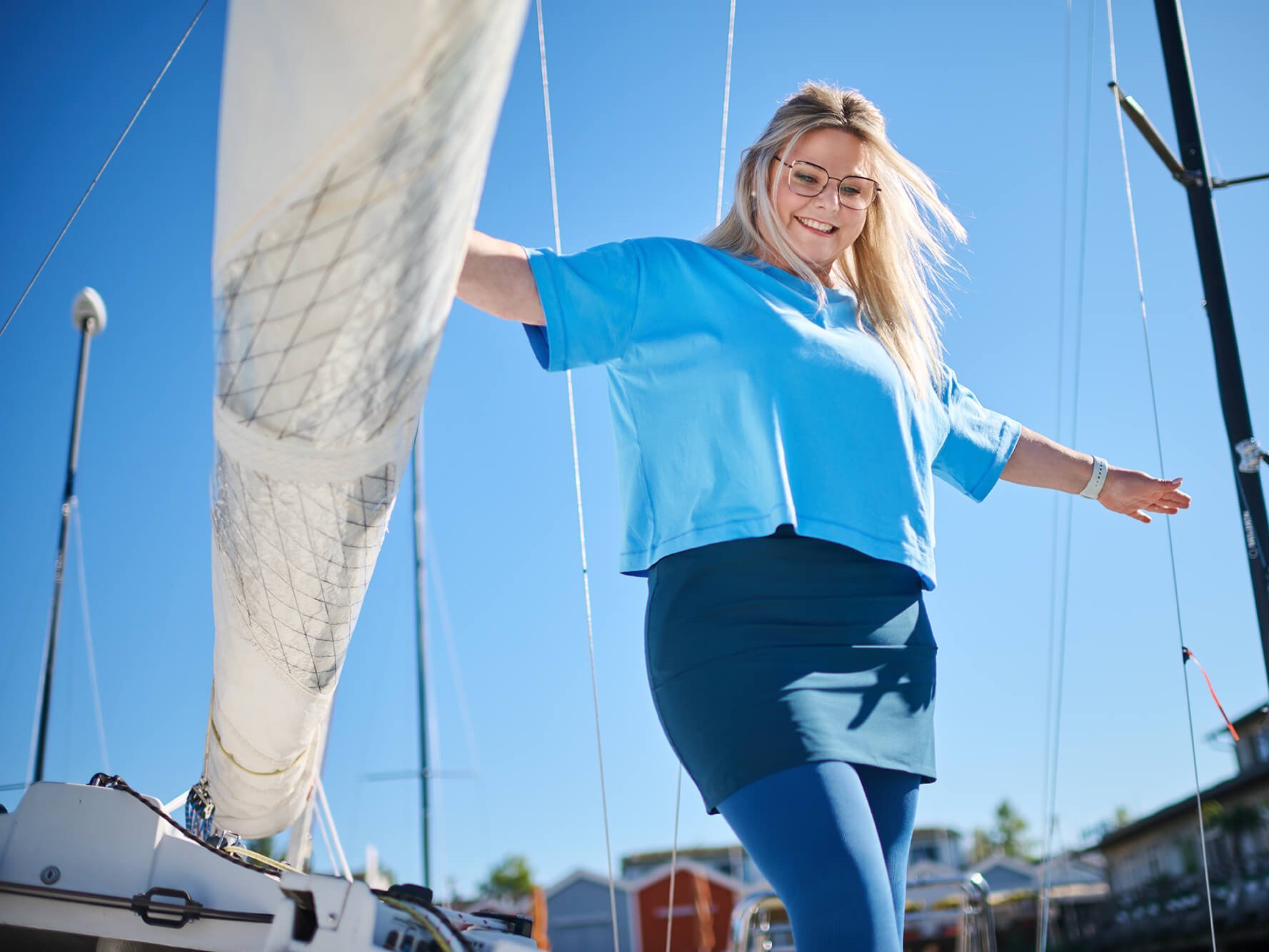 Frau in blauem T-Shirt und dunklem Rock und curaflow Lip-/Lymph-Versorgung von Bauerfeind steht mit ausgebreiteten Armen auf einem Segelboot bei klarem Himmel.