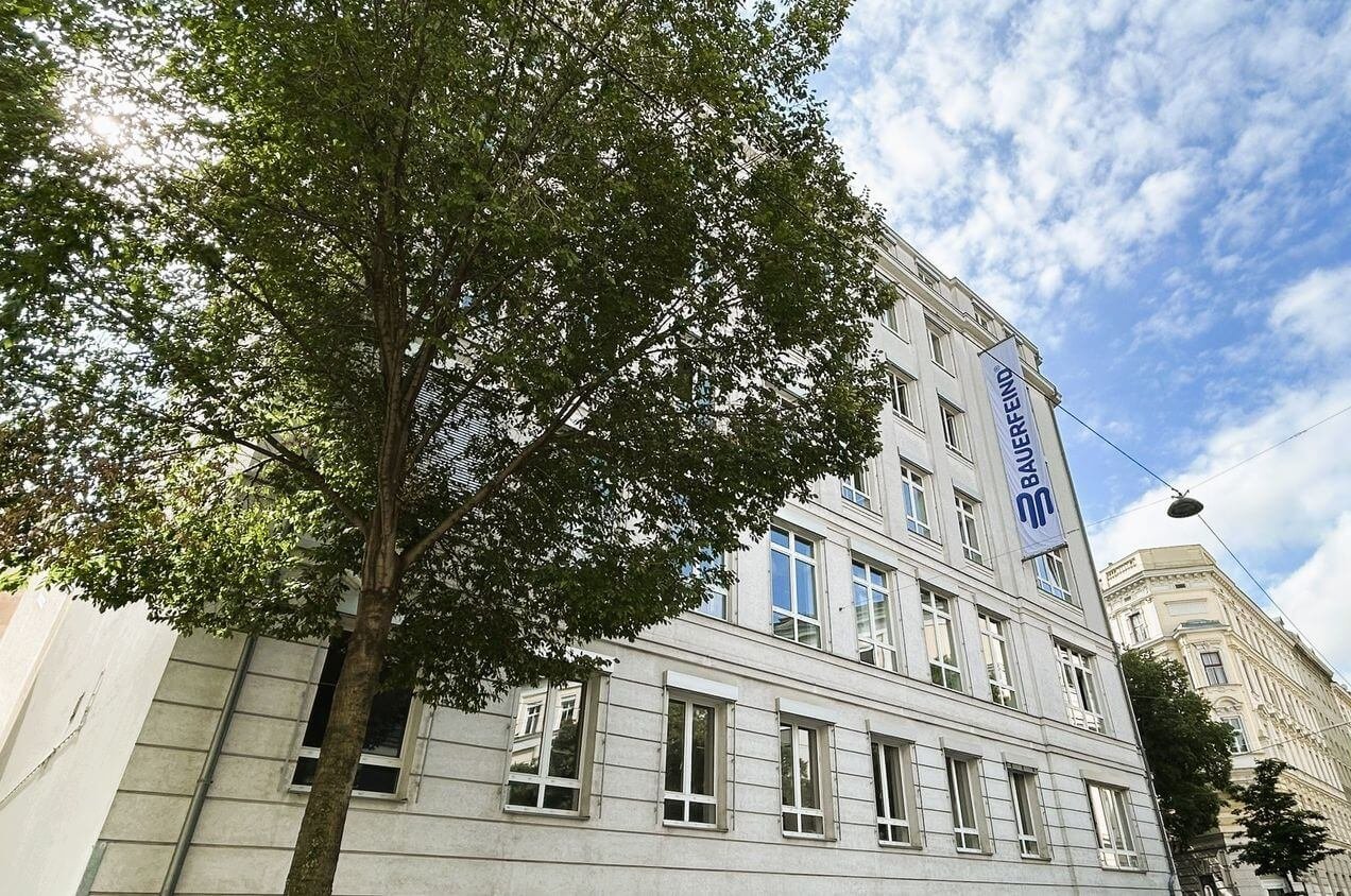 Strassenansicht eines mehrstöckigen, hellen Bürogebäudes mit dem Logo und Schriftzug „Bauerfeind“ auf einem vertikal angebrachten Banner. Im Vordergrund steht ein grosser Laubbaum, im Hintergrund sind weitere Altbauten zu sehen.