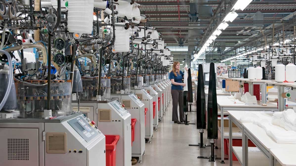 Innenansicht der Produktionshalle bei Bauerfeind: Mitarbeiterinnen und Mitarbeiter bedienen Maschinen und Anlagen, mehrere Arbeitstische sind mit Werkzeugen und Materialien bestückt.