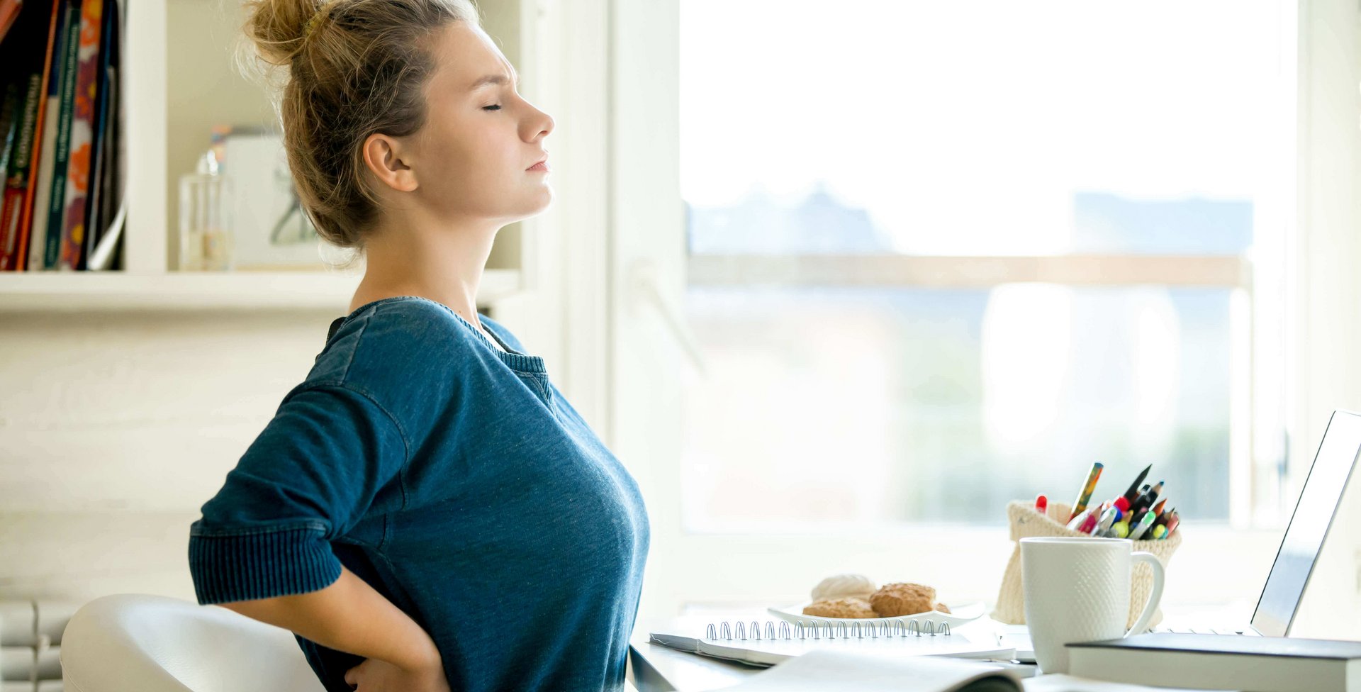 Frau mit Rückenbandage am Schreibtisch Eine Frau streckt sich an ihrem Homeoffice Arbeitsplatz im Sitzen.