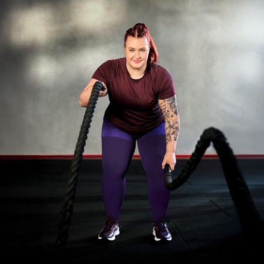 Eine Frau trainiert mit Battle Ropes vor einem neutralen, leicht beleuchteten Hintergrund. Sie trägt ein bordeauxfarbenes T-Shirt, violette Leggings und Sportschuhe. Ihr rechter Arm ist grossflächig tätowiert, und sie schaut fokussiert in die Kamera.