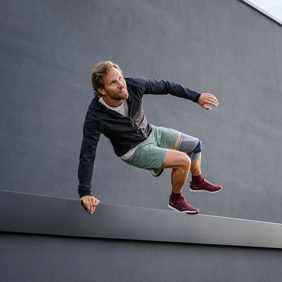 Abgebildet ist ein sportlicher Mann, der über ein Geländer vor einem Gebäude springt. Er trägt eine GenuTrian, eine schmerzlindernde Kniegelenksbandage von Bauerfeind, am linken Knie.