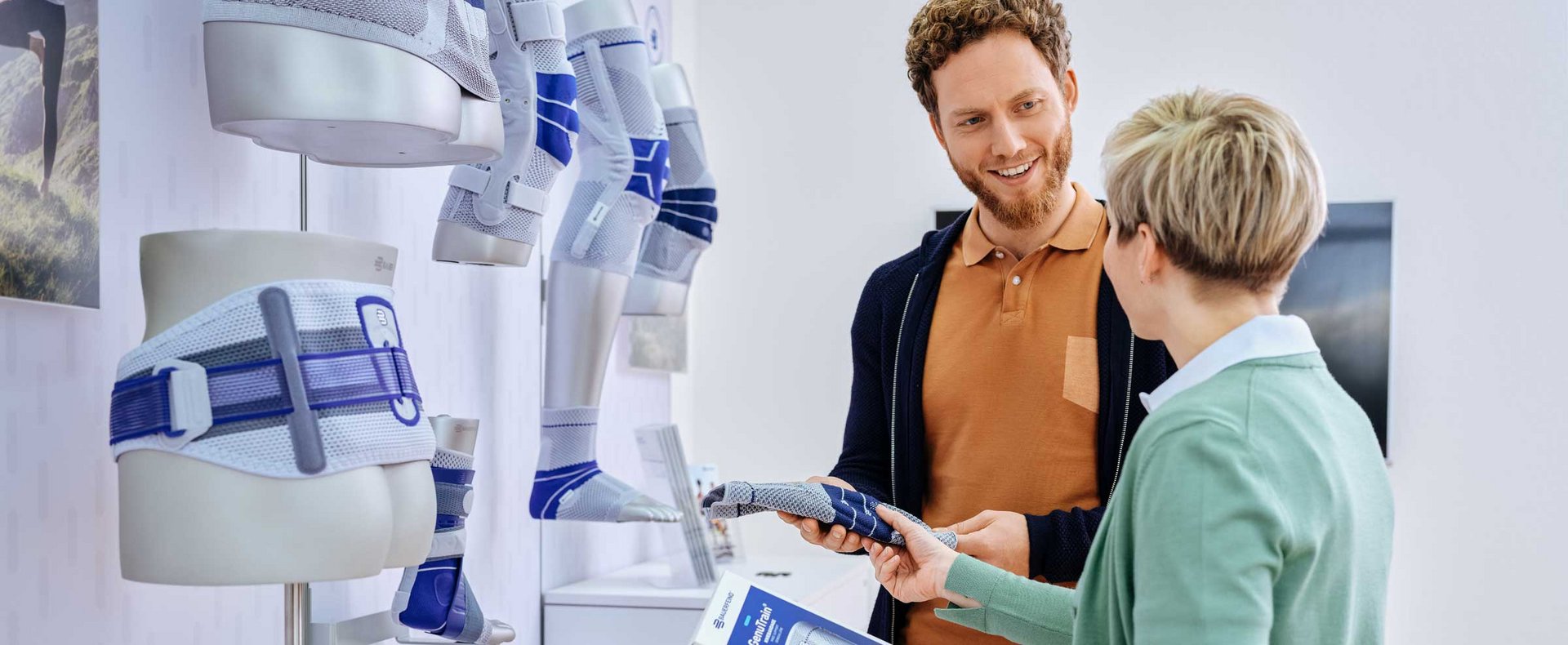 Beratungsgespräch zu Bandagen: Eine Person zeigt einem Hilfesuchenden eine Bandage und erklärt deren Anwendung. Im Hintergrund sind Beratungselemente wie Informationsmaterialien oder Displayflächen erkennbar.