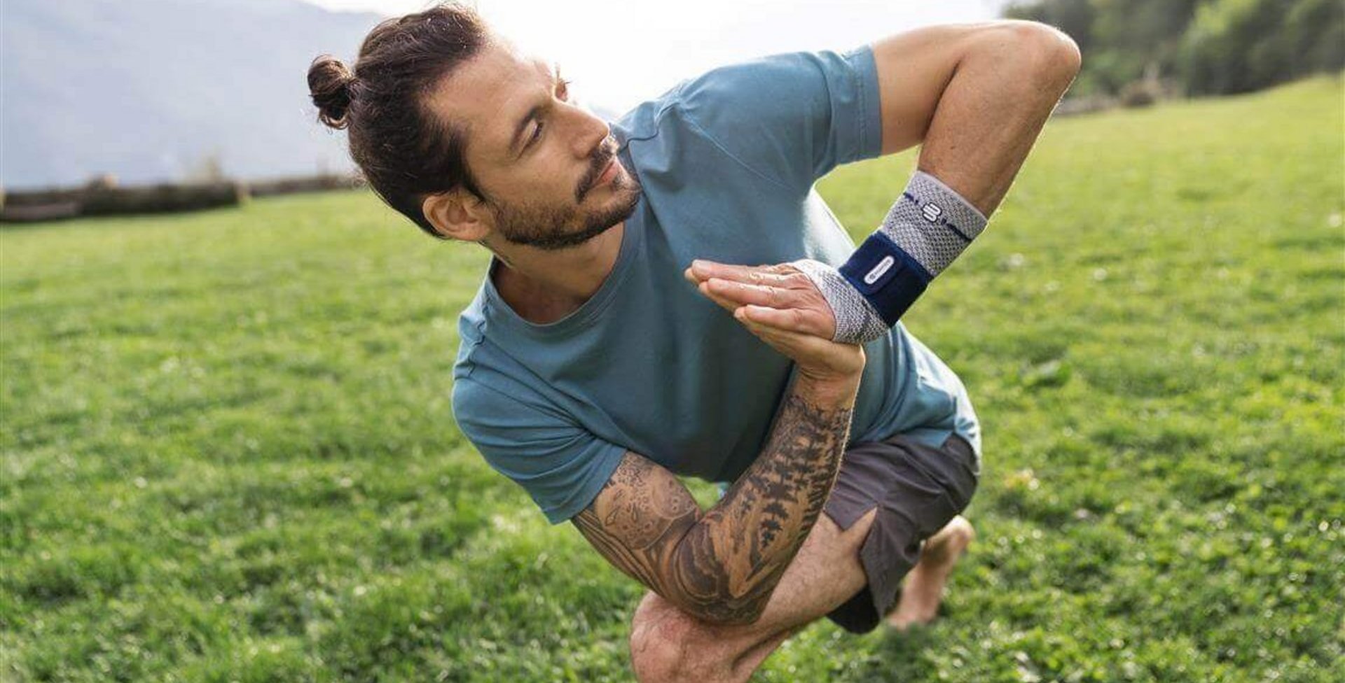 Abgebildet ist ein junger Mann auf einer Wiese in Yoga-Pose. Zur Unterstützung seines Handgelenks trägt er eine ManuTrain Handgelenkbandage von Bauerfeind.