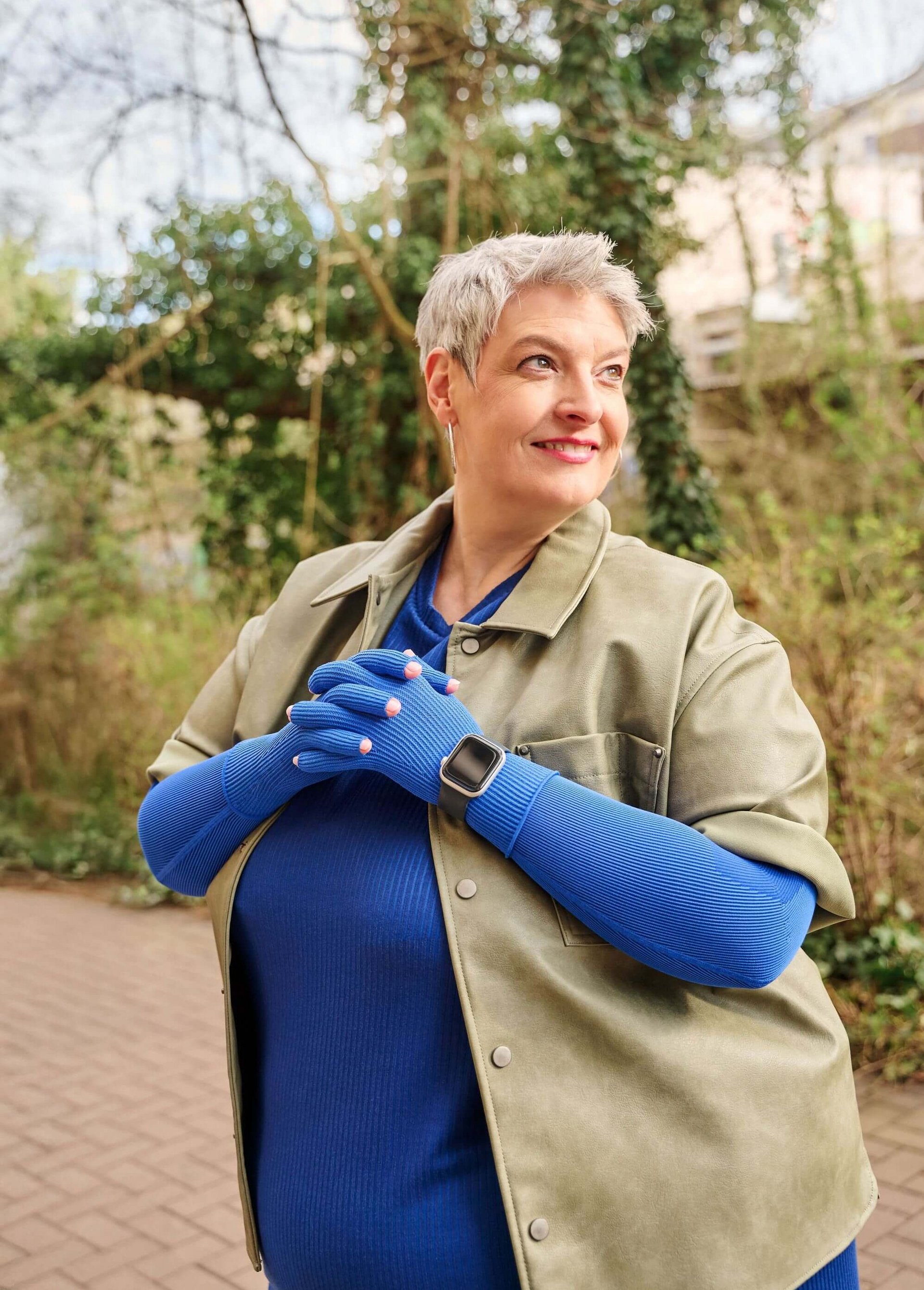 Ältere Frau mit kurzen grauen Haaren steht draussen auf einem gepflasterten Weg und lächelt zur Seite. Sie trägt ein blaues Oberteil mit dazu passenden blauen Handschuhen, eine olivgrüne Jacke sowie eine Smartwatch am linken Handgelenk. Im Hintergrund sind Bäume und Kletterpflanzen zu sehen.