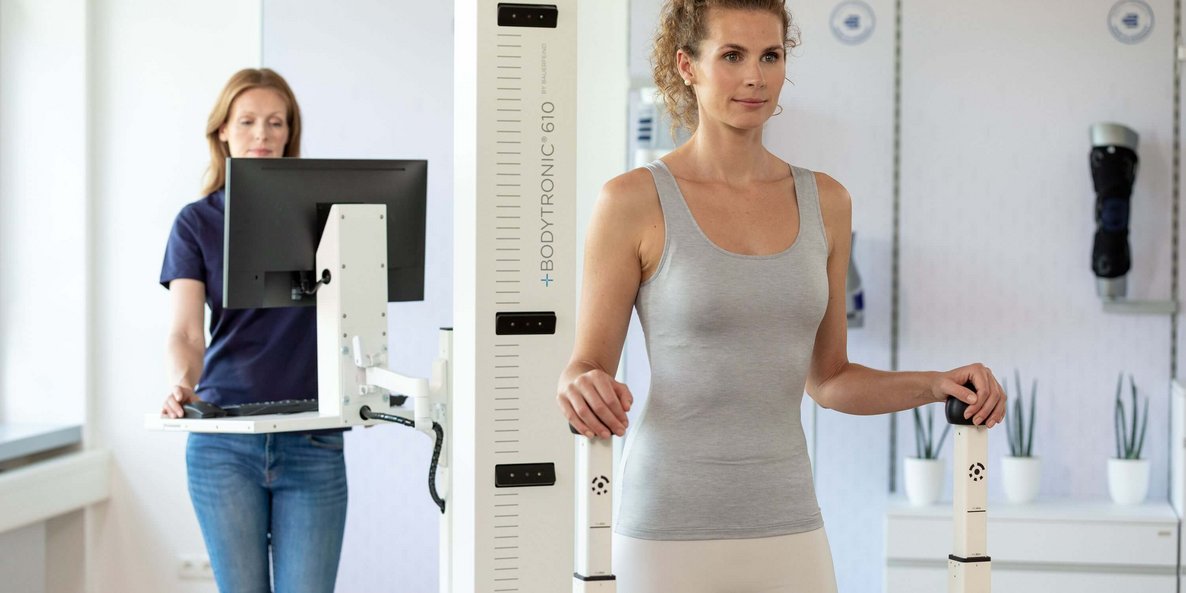 Frau in Sportbekleidung steht aufrecht an einem Bodytronic-610-Messgerät, das zur digitalen Körpervermessung dient. Im Hintergrund bedient eine Mitarbeiterin in Jeans und dunkelblauem Shirt einen Computer.