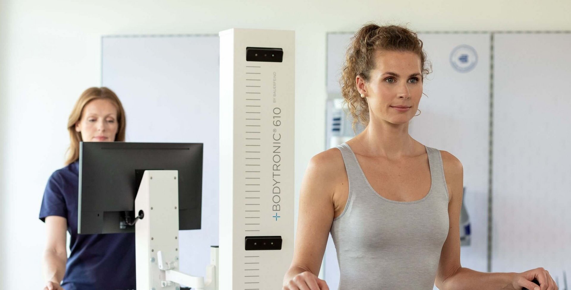 Frau in Sportbekleidung steht aufrecht an einem Bodytronic-610-Messgerät, das zur digitalen Körpervermessung dient. Im Hintergrund bedient eine Mitarbeiterin in Jeans und dunkelblauem Shirt einen Computer.