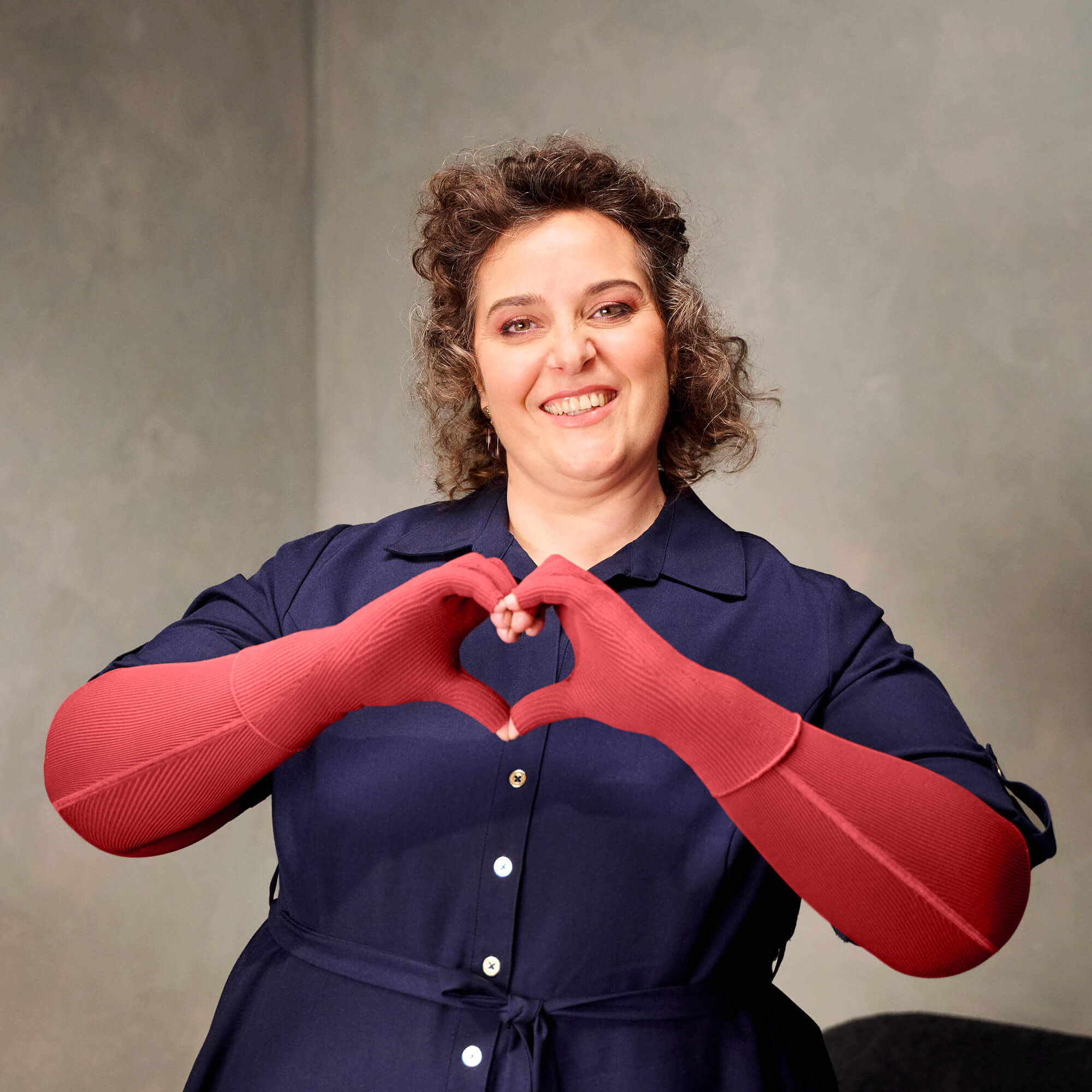 Eine lächelnde Frau steht vor einer hellgrauen Wand und formt mit ihren roten Kompressionshandschuhen ein Herz vor ihrer Brust.