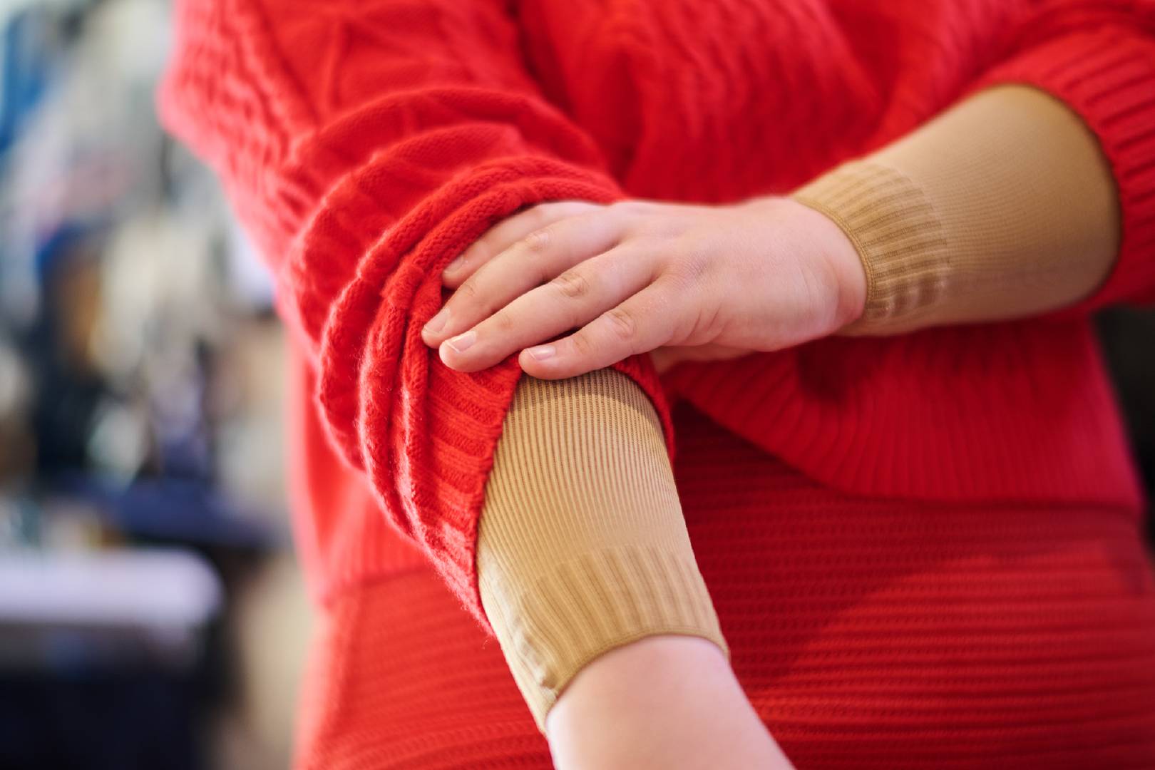 Nahaufnahme von einer Person, die den Ärmel ihres roten Strickpullovers hochzieht und einen beige-farbenen Kompressionsärmel darunter zeigt. Die Szene zeigt den Fokus auf die Unterstützung durch Kompressionskleidung im Alltag.